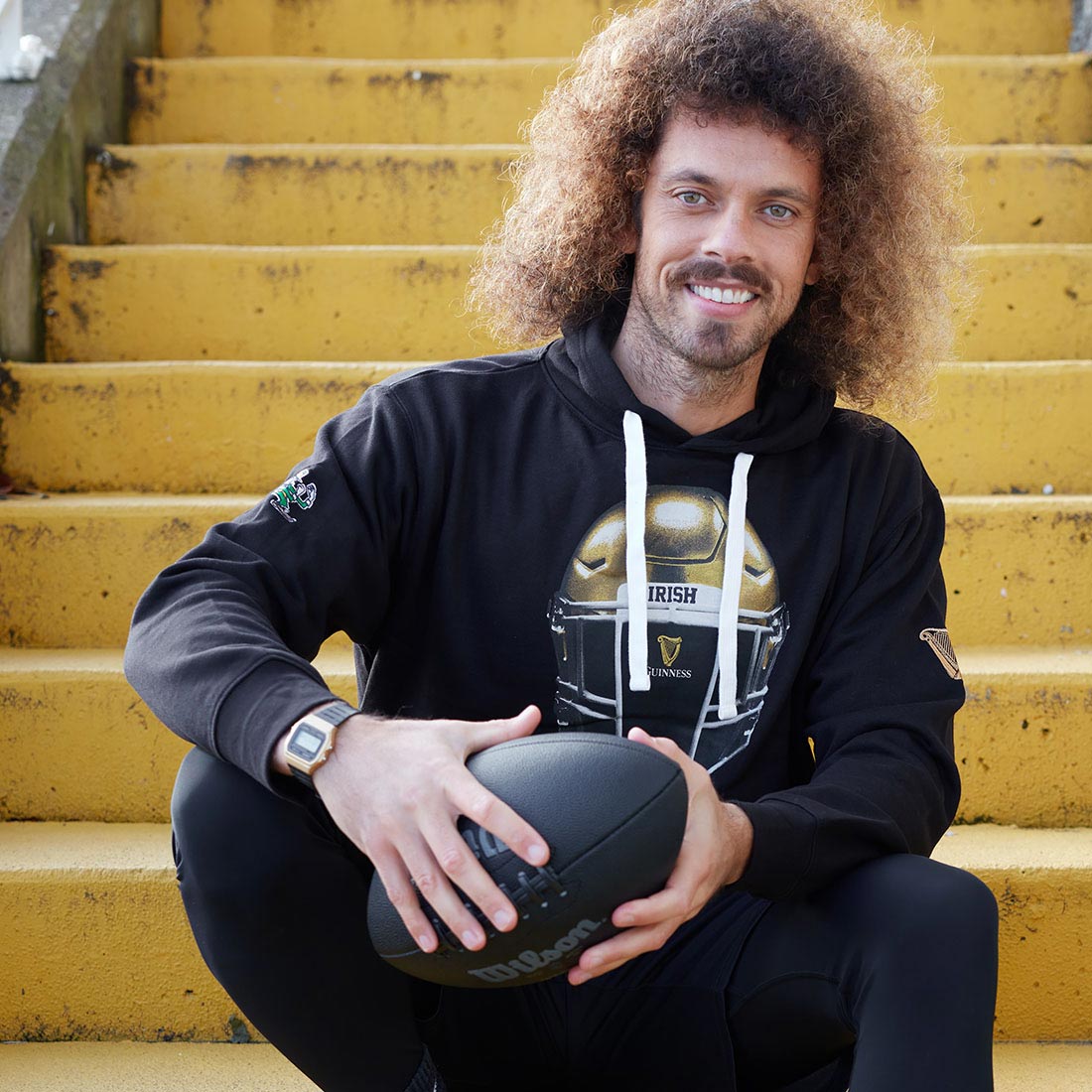 A man with curly hair sitting on yellow steps comfortably holding a Guinness Notre Dame Helmet Hoodie Black.