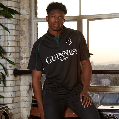 A man in a Guinness Black & Green Short Sleeve Rugby Jersey sitting on top of a barrel.