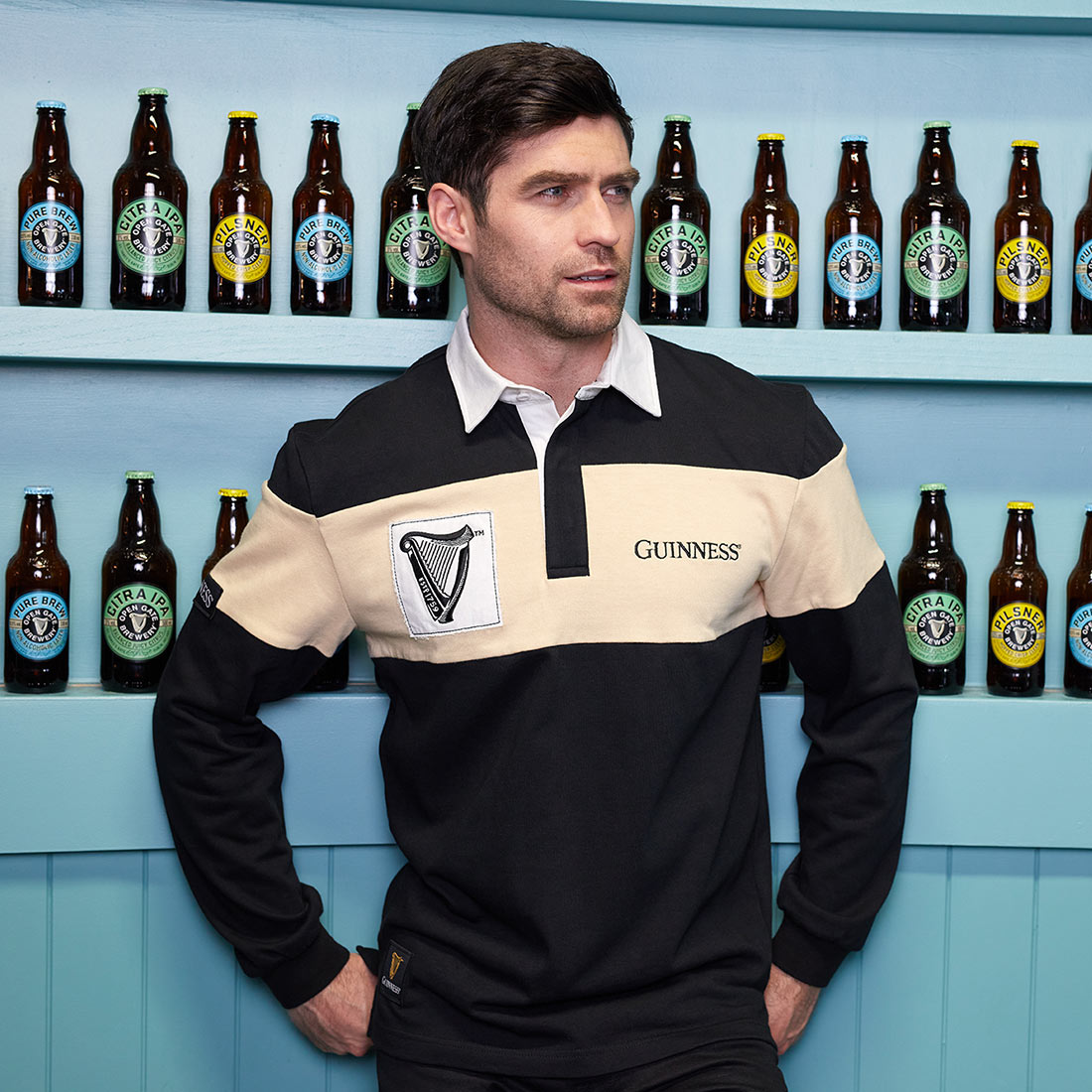 A man, wearing a Traditional Rugby Jersey with Cream panel and Harp logo patch, is leaning against a wall of beer bottles with prominent Guinness branding and the Harp logo.