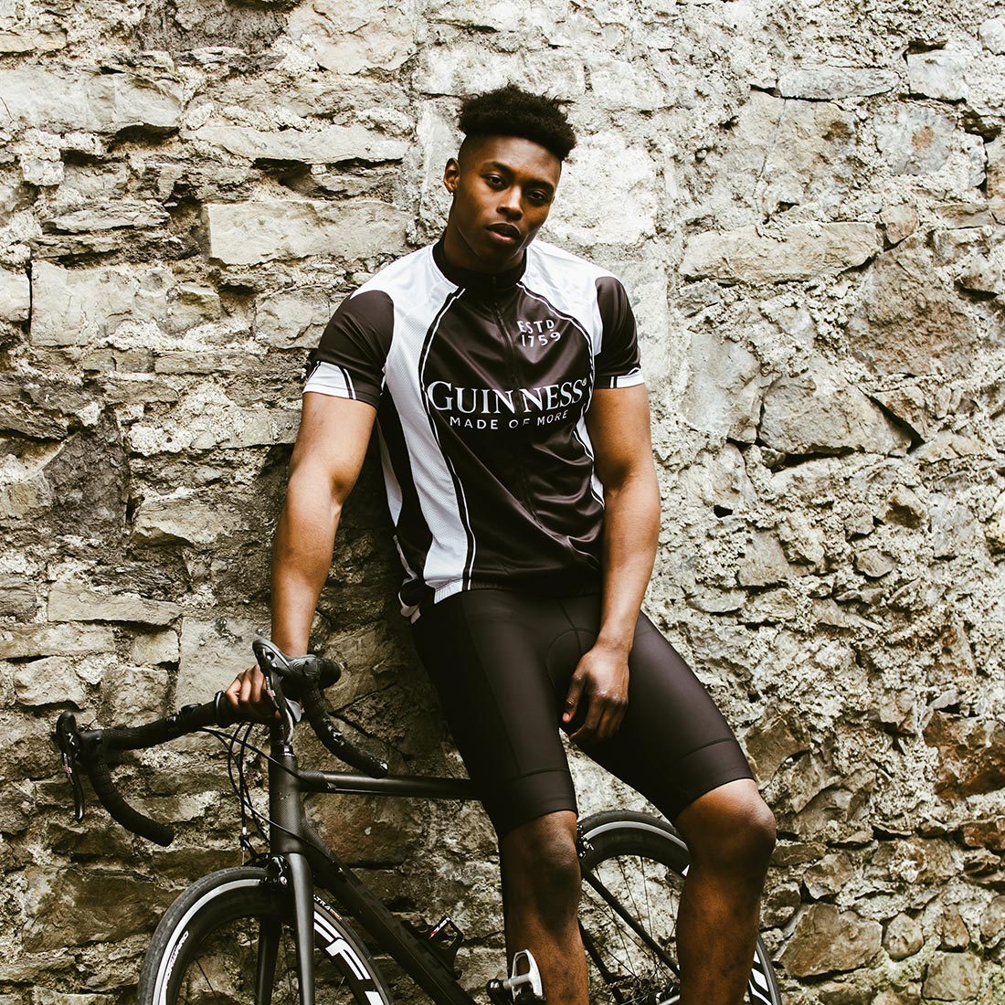 A man wearing a Guinness Performance Cycling Jersey leans against a stone wall with a bicycle.