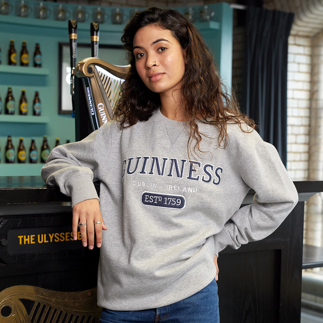 A woman is leaning against a bar in Dublin, Ireland, wearing a Guinness Grey Crew Neck Sweatshirt.