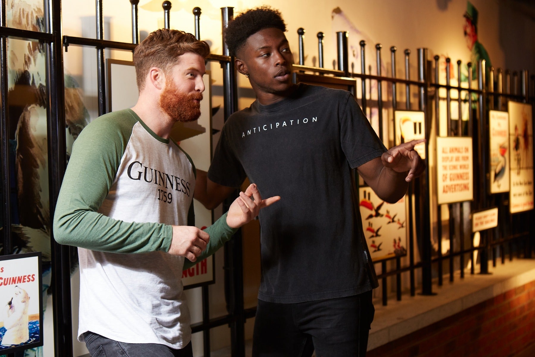 Two men stand outside by a black fence, each pointing the same way. One wears a Guinness Premium Vintage Tortoise Back Graphic Tee, while the other sports a black shirt with "Anticipation" in a distressed effect.