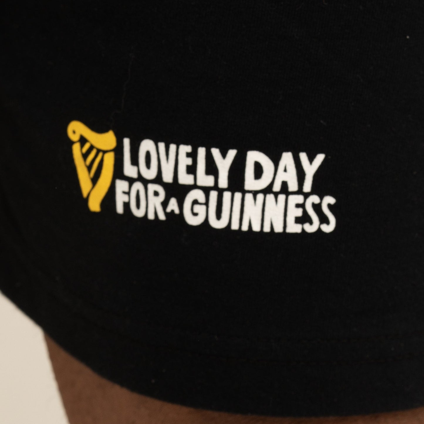 The Guinness Seb Curi Beach Short - Black from Guinness Webstore US features black fabric with white and yellow "Lovely Day for a Guinness" text and a harp logo, inspired by the Sebastian Curi collaboration.