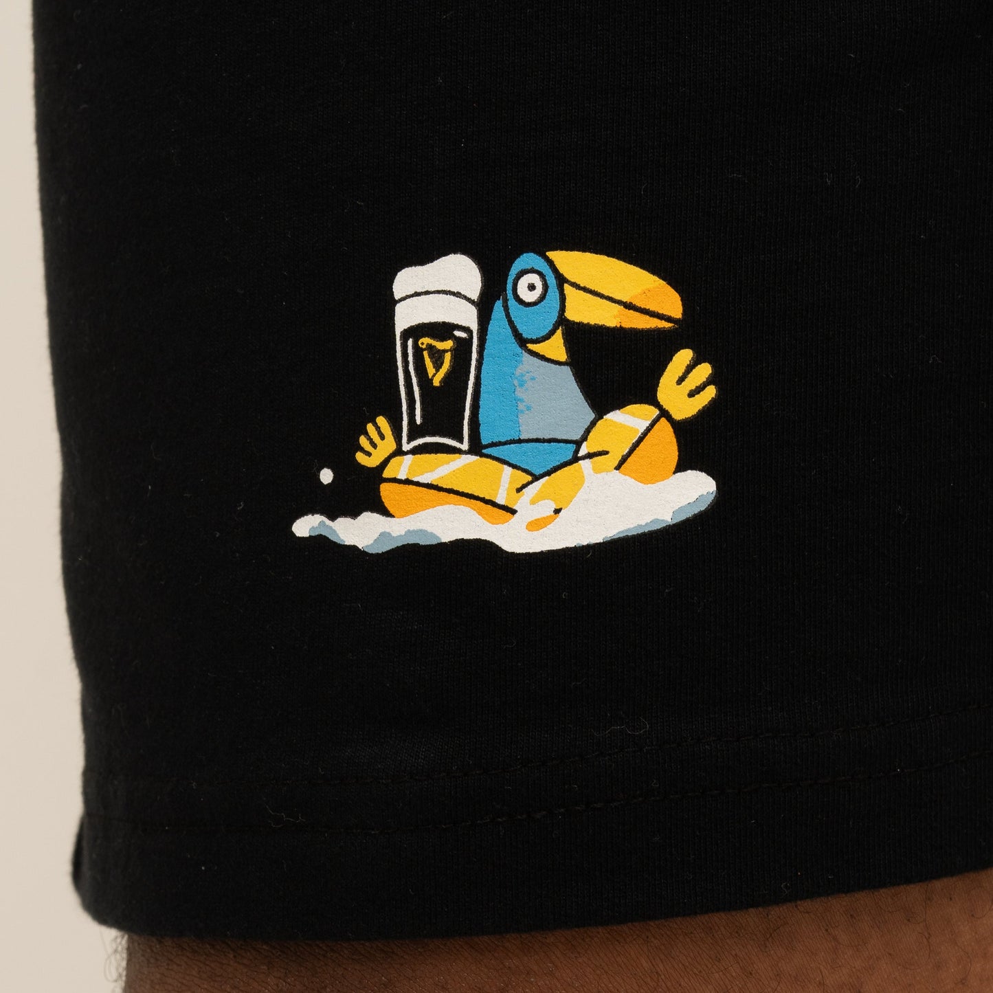The Guinness Seb Curi Beach Short - Black from Guinness Webstore US features a Sebastian Curi-inspired blue and yellow toucan sitting in foam beside a pint of dark beer, illustrated and printed on black fabric.