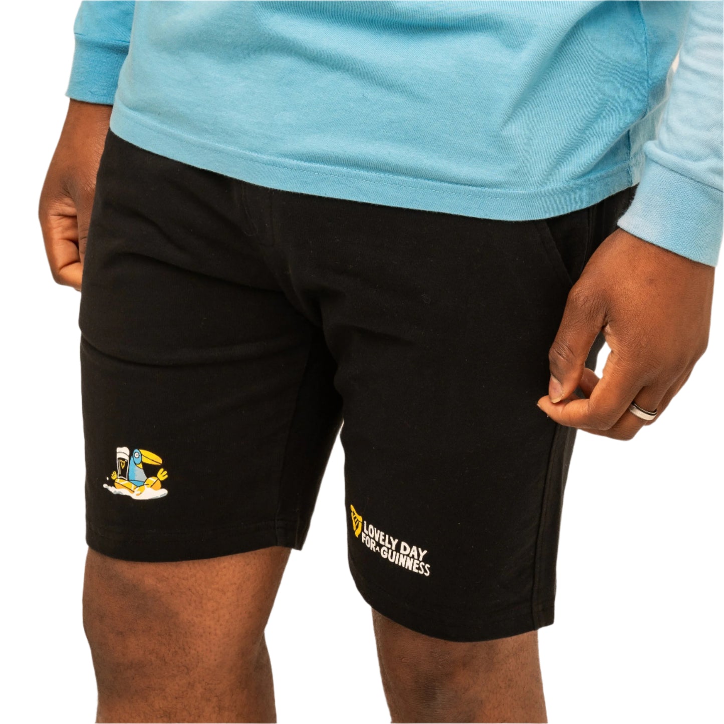 A person wearing a light blue long-sleeve shirt and black Guinness Seb Curi Beach Short from Guinness Webstore US, featuring small graphic prints and text on the left leg from the Sebastian Curi collaboration.