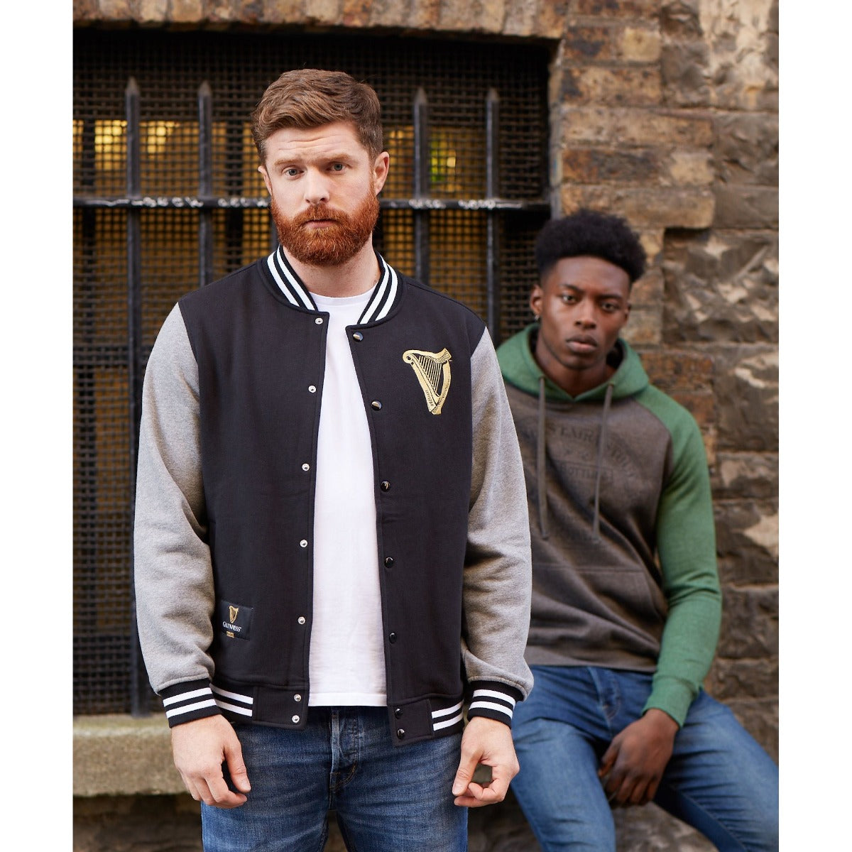 A man and a woman wearing Guinness varsity jackets standing next to a brick wall.