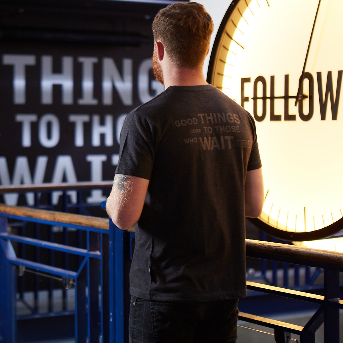 A man wearing a Guinness Surf Tee, a black t-shirt made with durable cotton blend fabric, looking at a clock.
