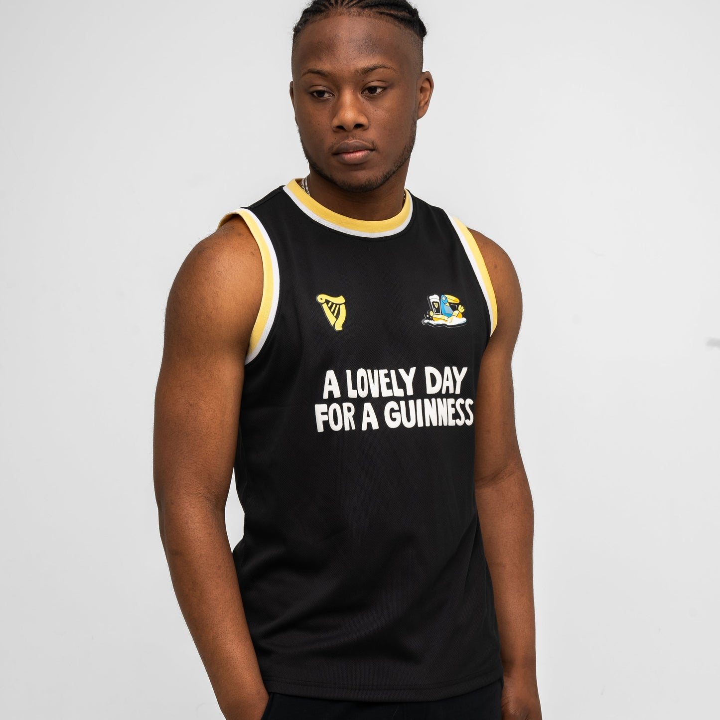 A man wears a black Guinness Seb Curi Basketball Jersey from Guinness Webstore US, featuring yellow and white trim and the slogan “A LOVELY DAY FOR A GUINNESS,” standing against a plain light background.