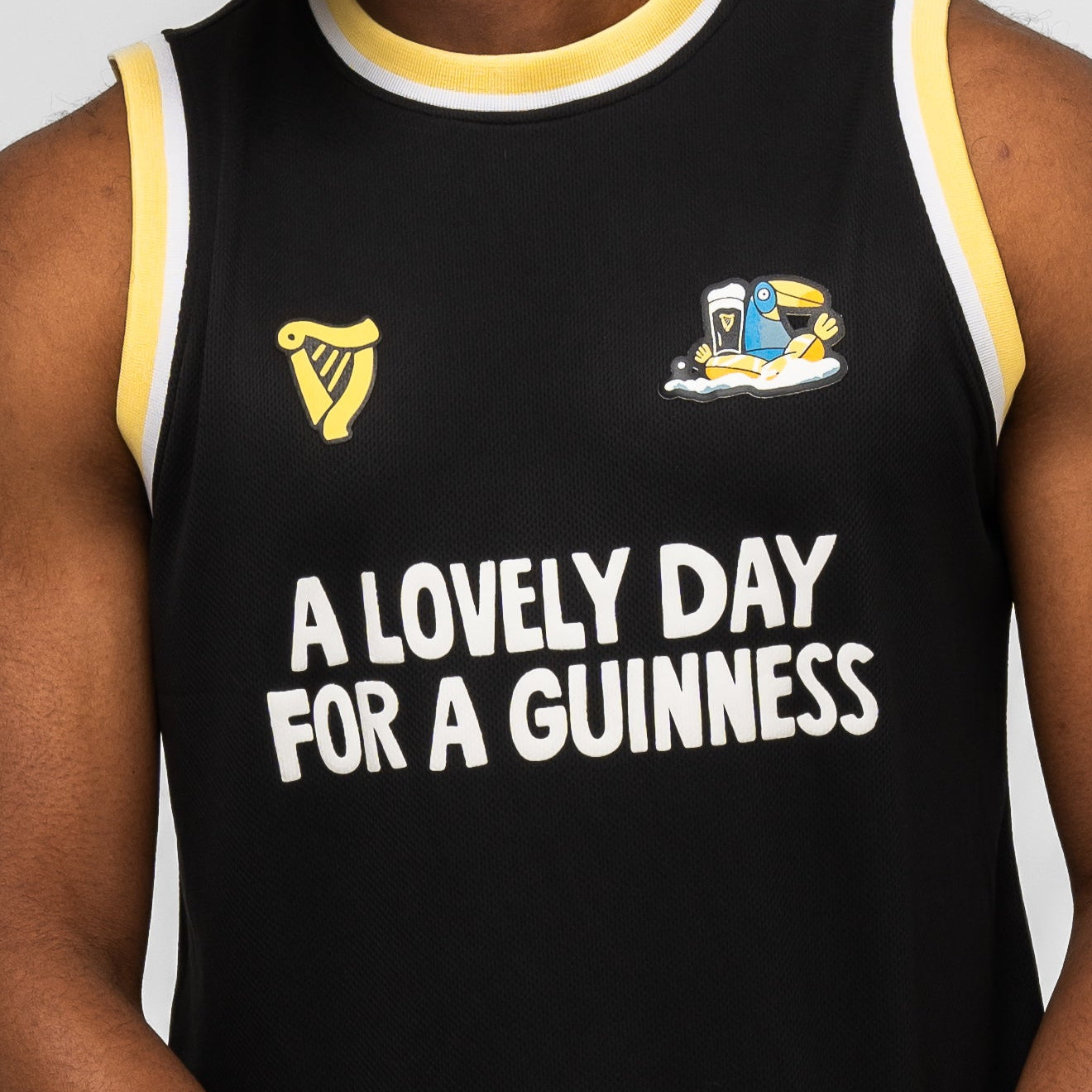 A person wears the Guinness Seb Curi Basketball Jersey from Guinness Webstore US—a black sleeveless top with yellow trim, "A LOVELY DAY FOR A GUINNESS" text, Guinness logos, and a cartoon toucan in Sebastian Curi’s limited-edition style.