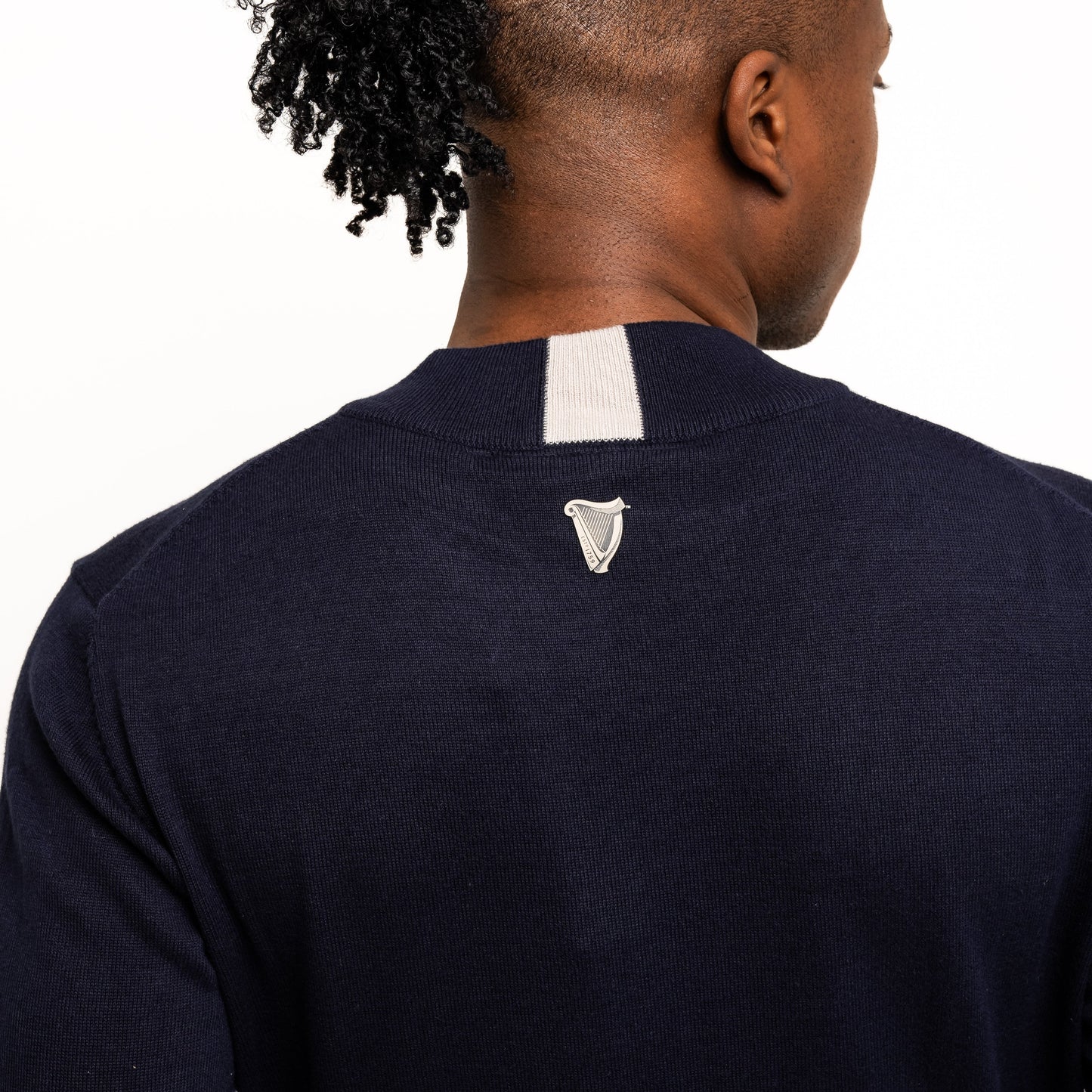 A person with short, braided hair pulled back is wearing the Guinness Performance Cotton Sweater - Navy from Guinness Webstore US, featuring moisture-wicking cotton, a small white patch, and an embroidered upper-back logo—ideal for golf.