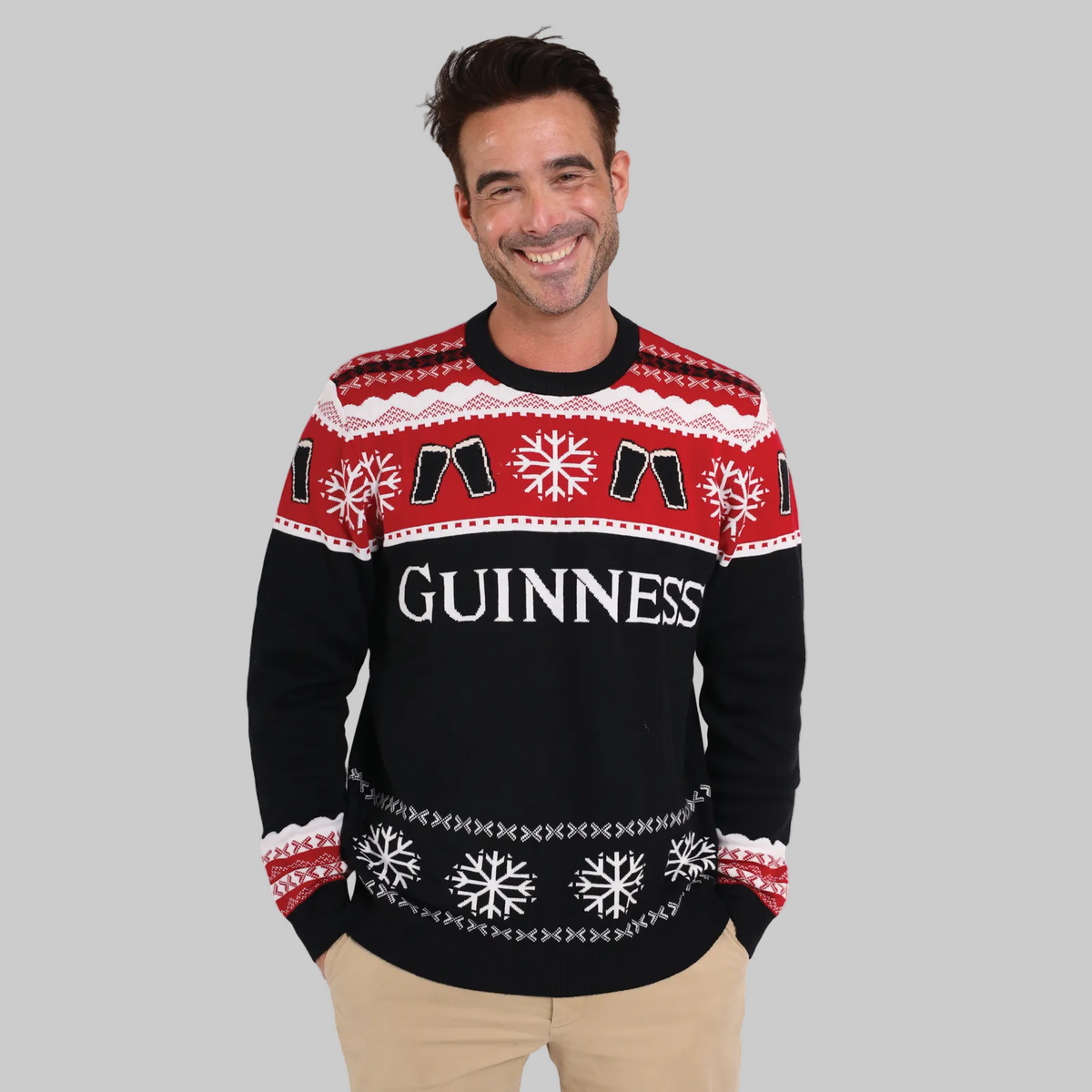 A man smiles with hands in pockets, wearing the Official Guinness Holiday Sweater—a festive red, black, and white knit featuring snowflake patterns and "Guinness" on the front—perfect for any holiday wardrobe.
