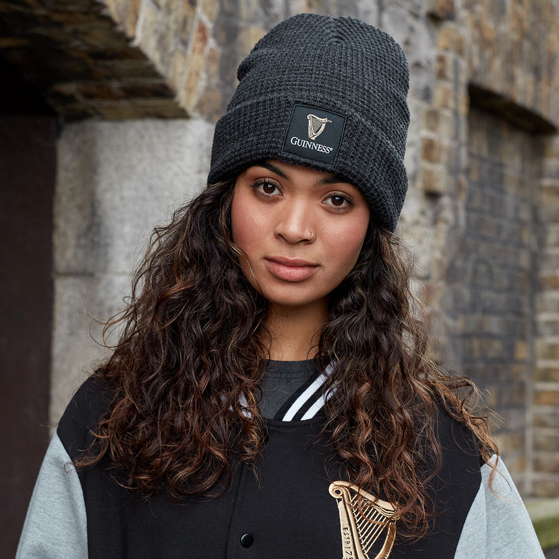 A woman with long curly hair wears the Guinness Thermal Warm Beanie by Guinness, paired with a matching jacket, standing in front of a brick wall.