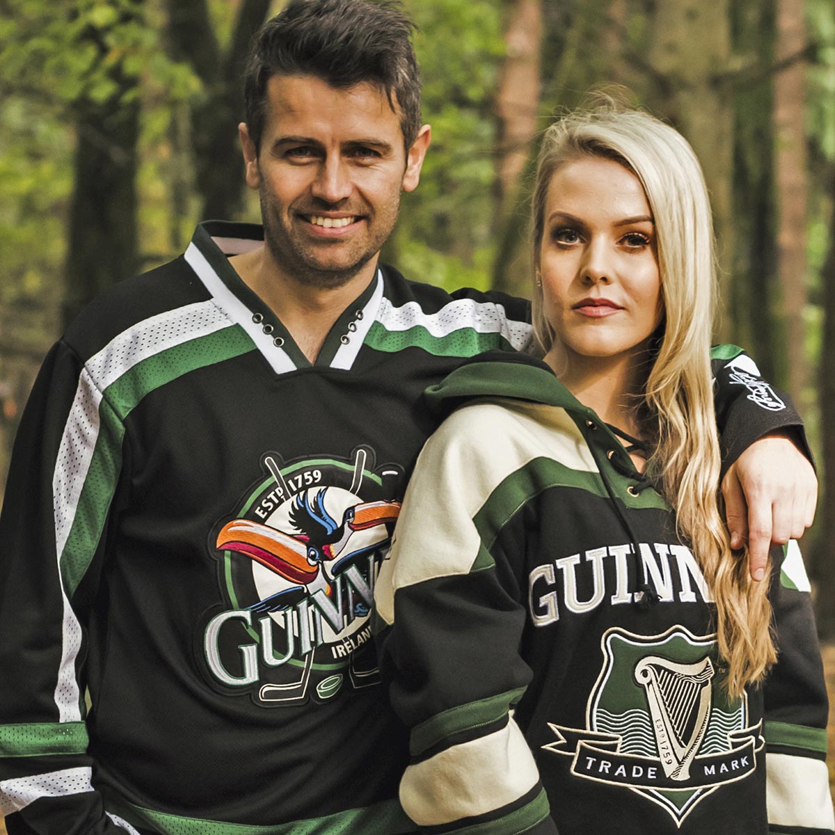 A man and woman wearing Guinness Toucan Hockey Jerseys Black and Green are posing for a photo in the woods.