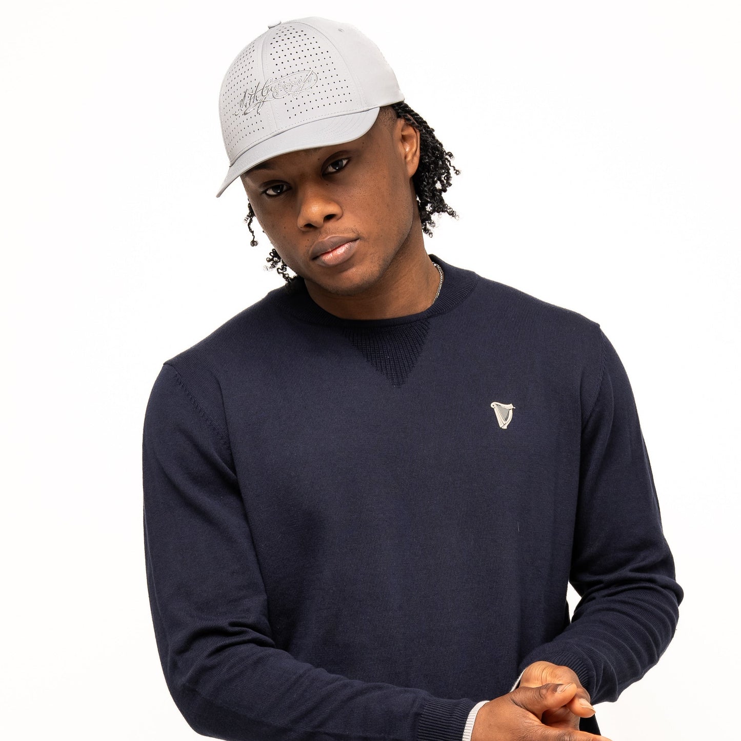 A person wears a light gray baseball cap, Guinness Webstore US Guinness Performance Cotton Sweater in navy, and blue pants, standing against a plain white background.