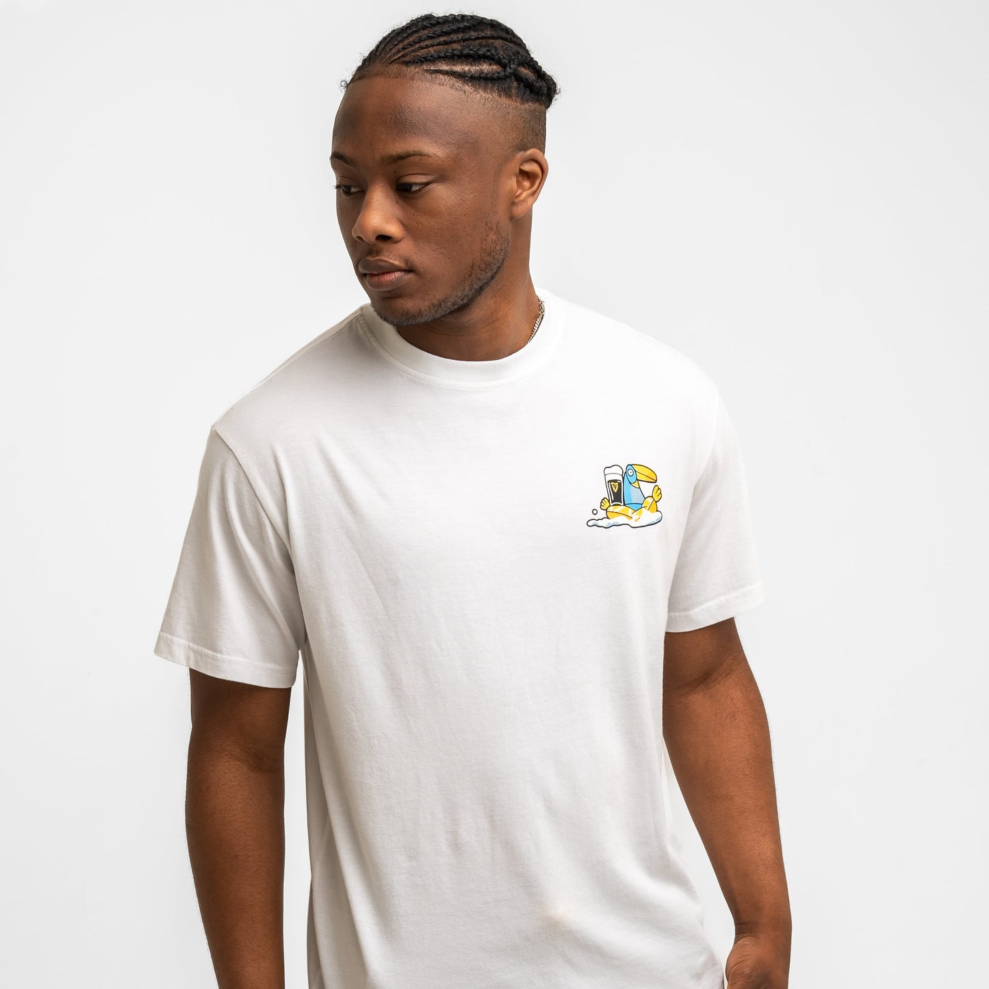 A man wears the Guinness Webstore US Guinness Seb Curi Oversize Tee - White, with a small, colorful left-chest graphic from the Sebastian Curi collab, standing against a plain white background and looking to his left. The tee has a small cartoon graphic of a blue toucan with a pint of Guinness on the chest