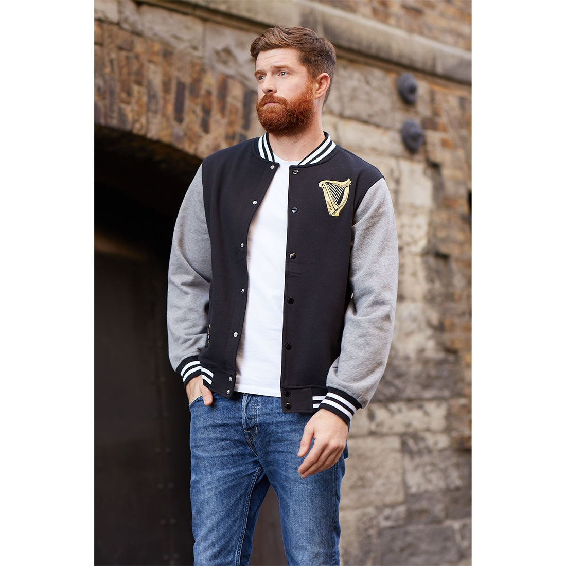 A man with a beard and jeans wearing a Guinness Letterman Jacket, proudly displaying Guinness merchandise.