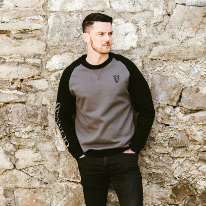 A man leaning against a stone wall wearing a mid-weight, unisex Guinness Long Sleeve Sweater.