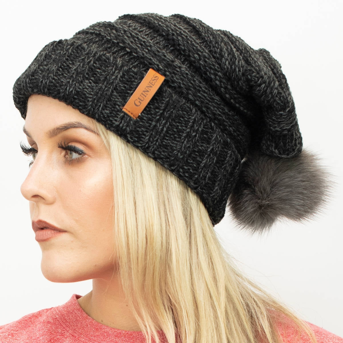 A woman wearing a Guinness Dark Grey Slouchy Bauble Beanie with Brown Leather Patch, perfect for the winter months.
