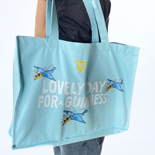 A person holds the Guinness Seb Curi Tote Bag – Blue from Guinness Webstore US, designed by Sebastian Curi with cartoon birds and "LOVELY DAY FOR A GUINNESS" text.