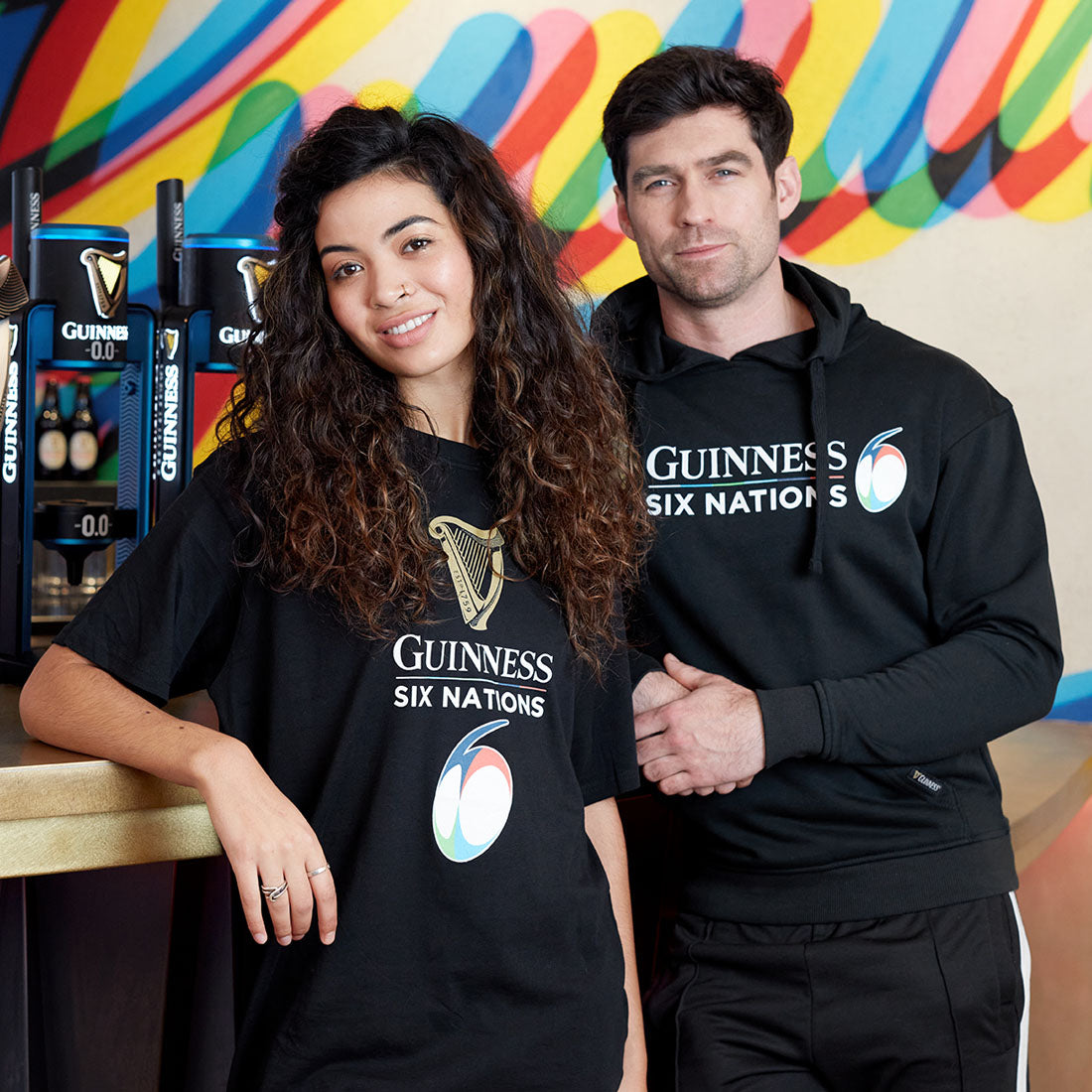 A man and a woman wearing Guinness hoodies stand in front of a colorful promotional backdrop.