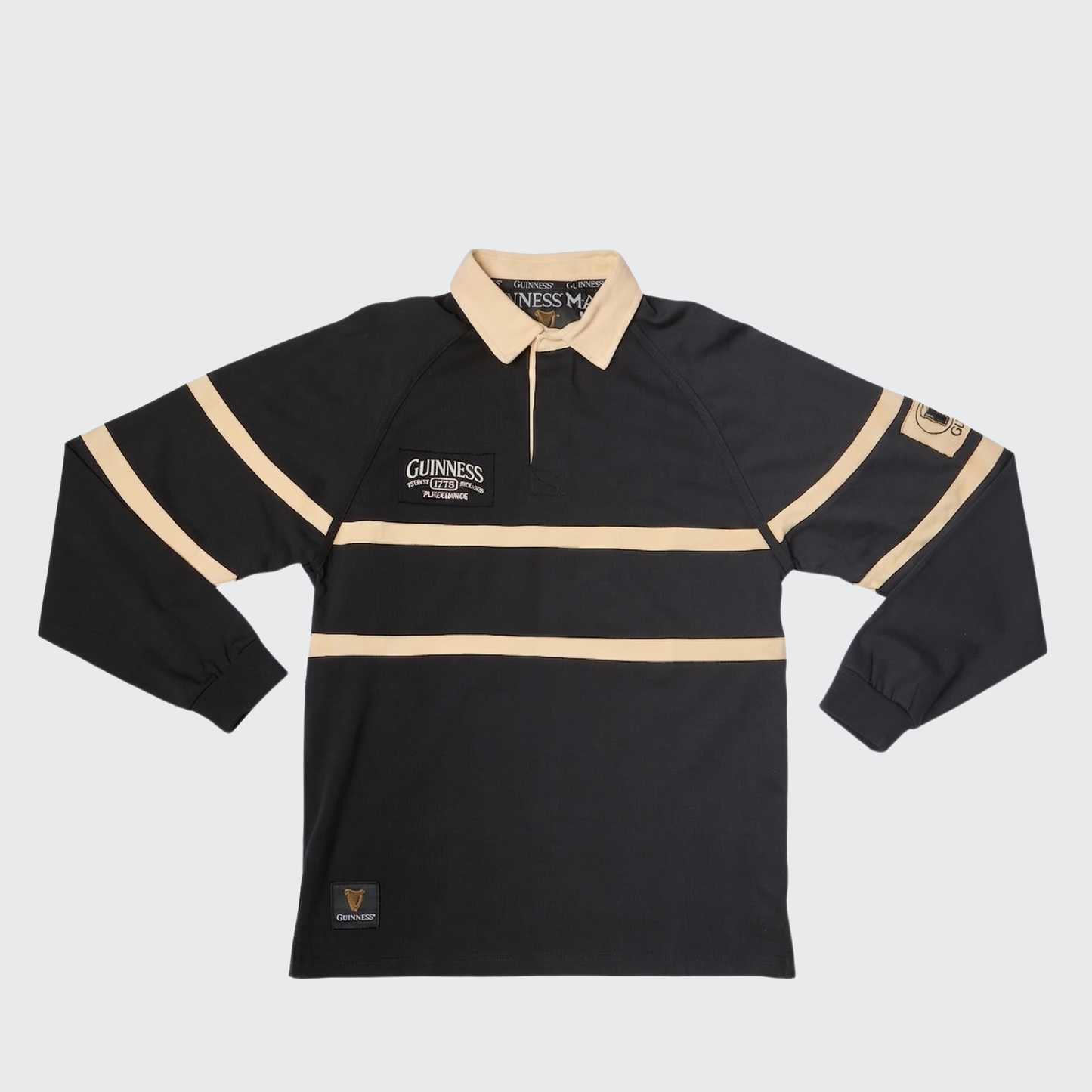 Guinness Black Traditional Rugby Jersey by Guinness, featuring a black long-sleeve design with tan collar and stripes, crafted from 100% cotton for lasting comfort. Includes Guinness branding on the chest and a sleeve patch, and is shown laid flat on a plain background.
