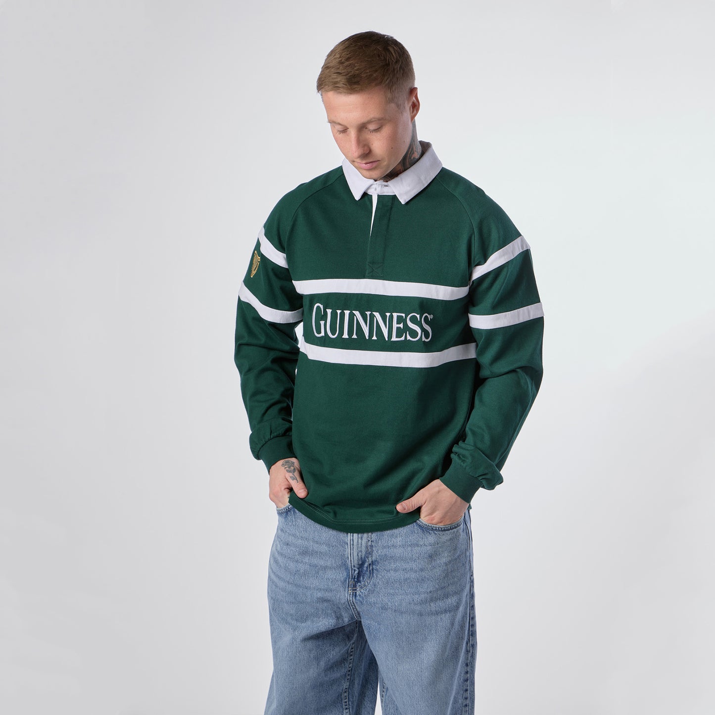 A person wearing the Guinness Green Traditional Rugby Jersey with light blue jeans stands against a plain white background, looking down.