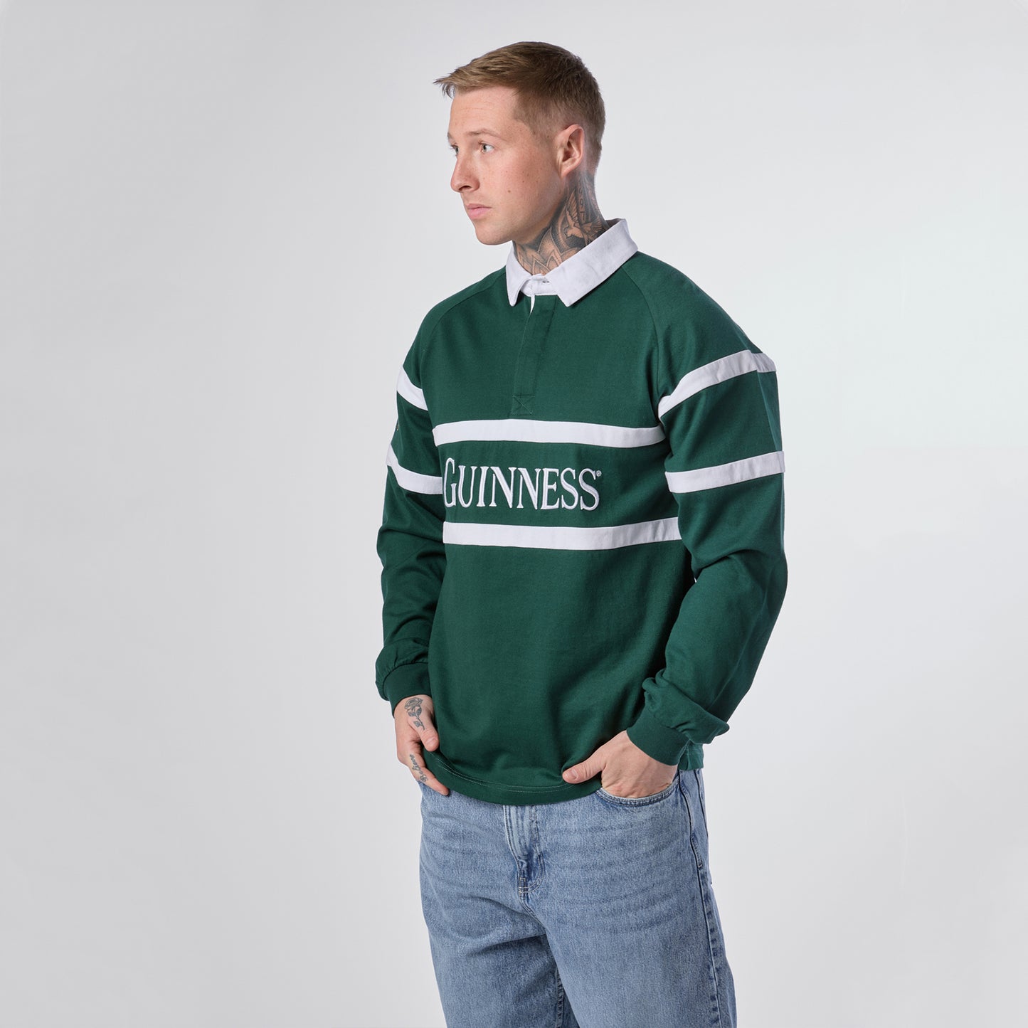 Wearing the Guinness Green Traditional Rugby Jersey by Guinness and blue jeans, a person stands against a plain light background, looking to the left.
