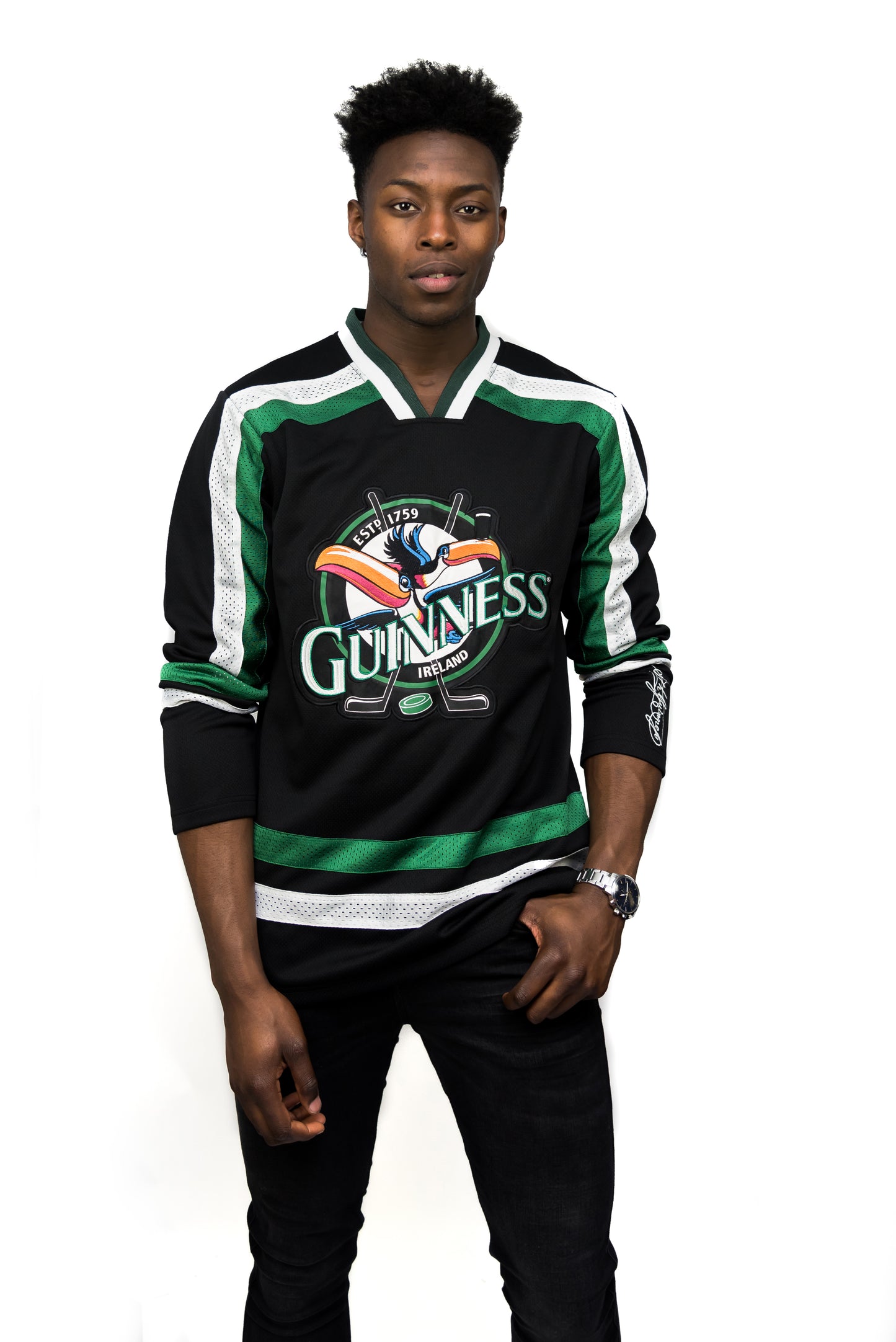 A man stands against a white background wearing the Guinness Toucan Hockey Jersey Black and Green by Guinness, paired with black pants.
