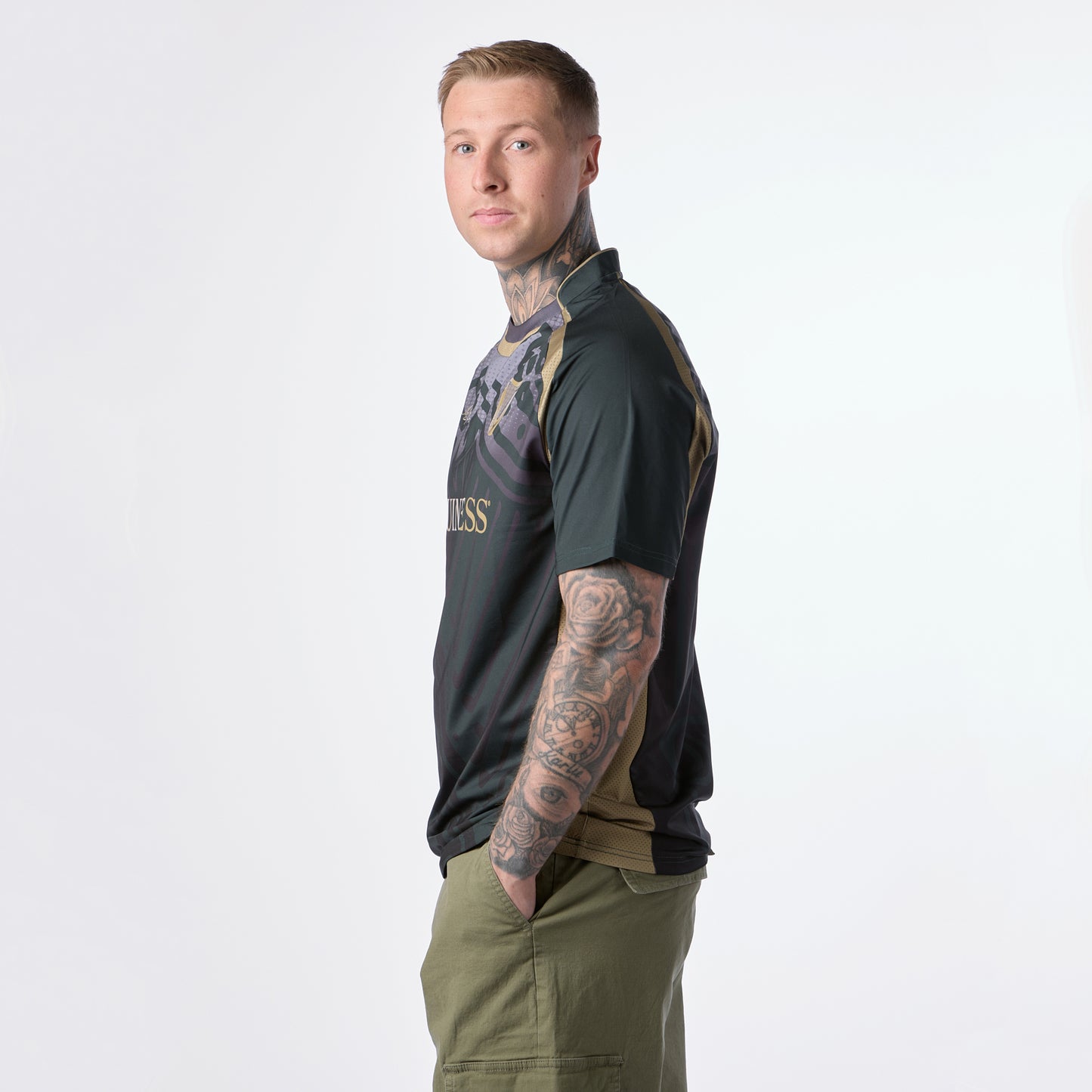 A person with tattoos on their arms stands in profile, wearing a Guinness Retro Harp Performance Soccer Jersey and olive green shorts, hands in pockets, against a plain light background.
