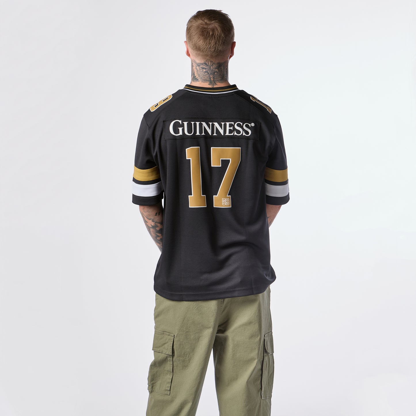 A person with tattoos is seen from behind wearing the Guinness Black American Football Jersey by Guinness Webstore US, featuring “GUINNESS” and the number 17, paired with green cargo pants.