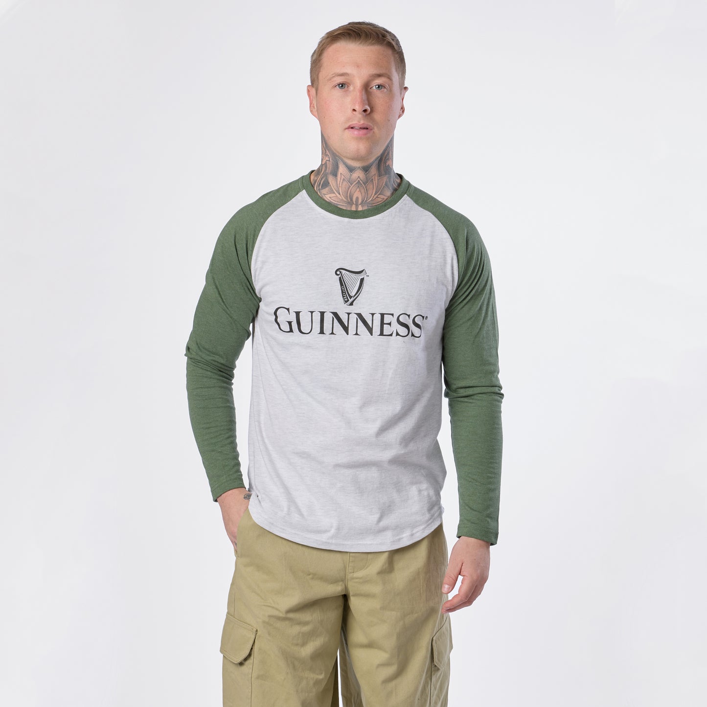 A man with tattoos wears the Guinness Baseball T-Shirt by Guinness in white and green along with khaki pants, standing against a plain white background and looking directly at the camera.