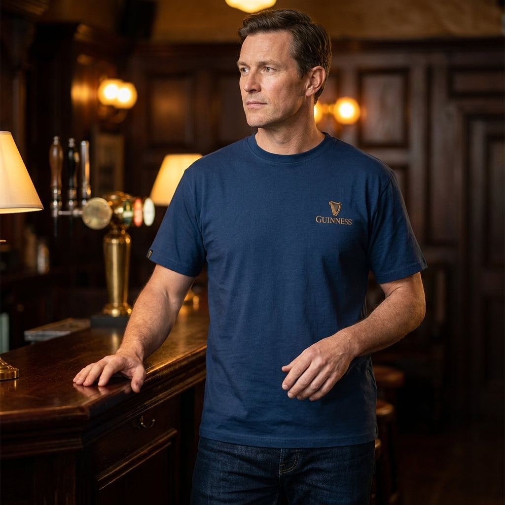 A man in a Guinness Gilroy Seal Tee – Blue from Guinness Webstore US stands at a wooden bar counter in a dimly lit pub with warm lighting and dark wood paneling.