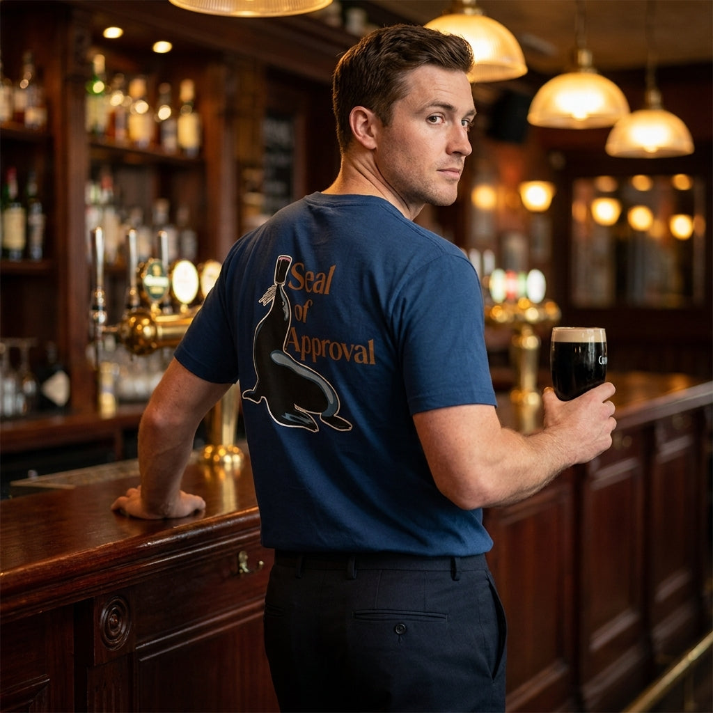 A man stands at a bar holding a pint of dark beer, wearing the Guinness Gilroy Seal Tee in blue from Guinness Webstore US, featuring a seal graphic with a pint of Guinness balanced on his nose and "Seal of Approval" on the back.
