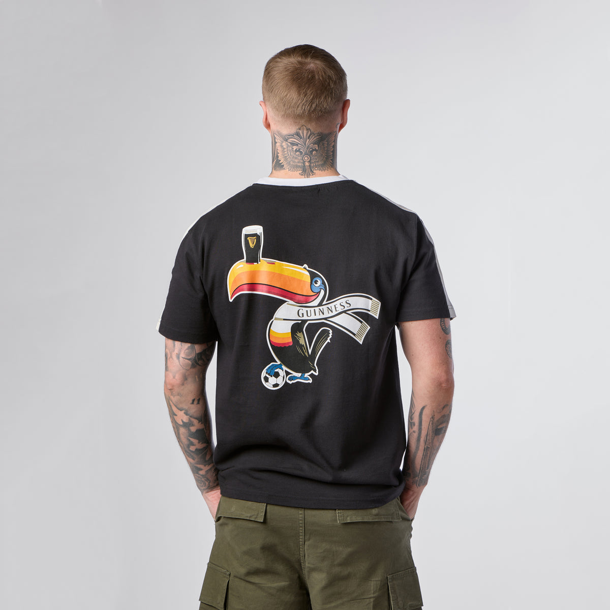 A person with tattoos on their neck and arms wears the Guinness Toucan Soccer Black Tee from Guinness Webstore US, which features a colorful toucan, a pint of Guinness, and a soccer ball graphic on the back.