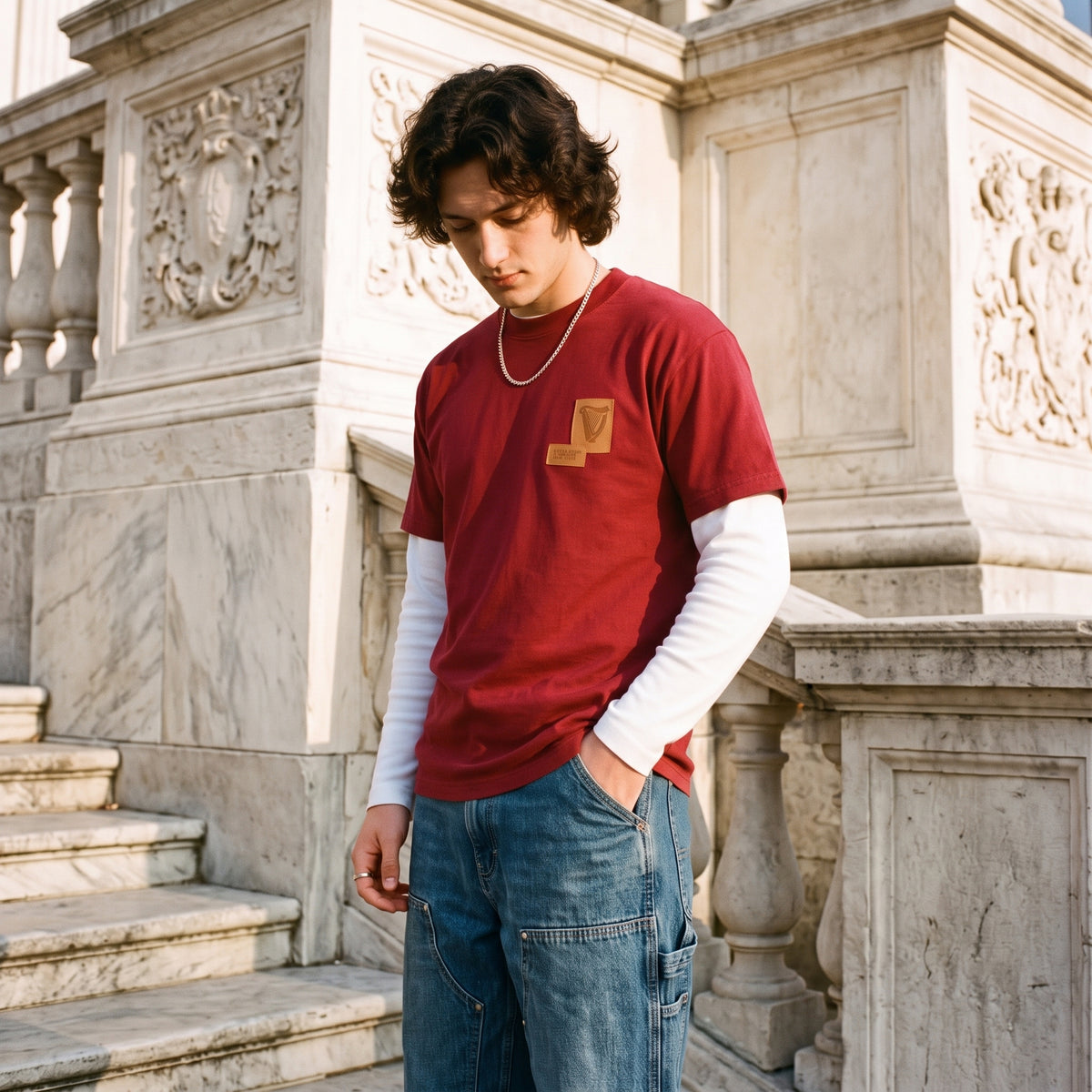 Wearing a Guinness Cooperage Burnt Red Heavyweight Tee over a white long-sleeve and blue jeans, someone stands on marble stairs with carved stone railings, looking down.