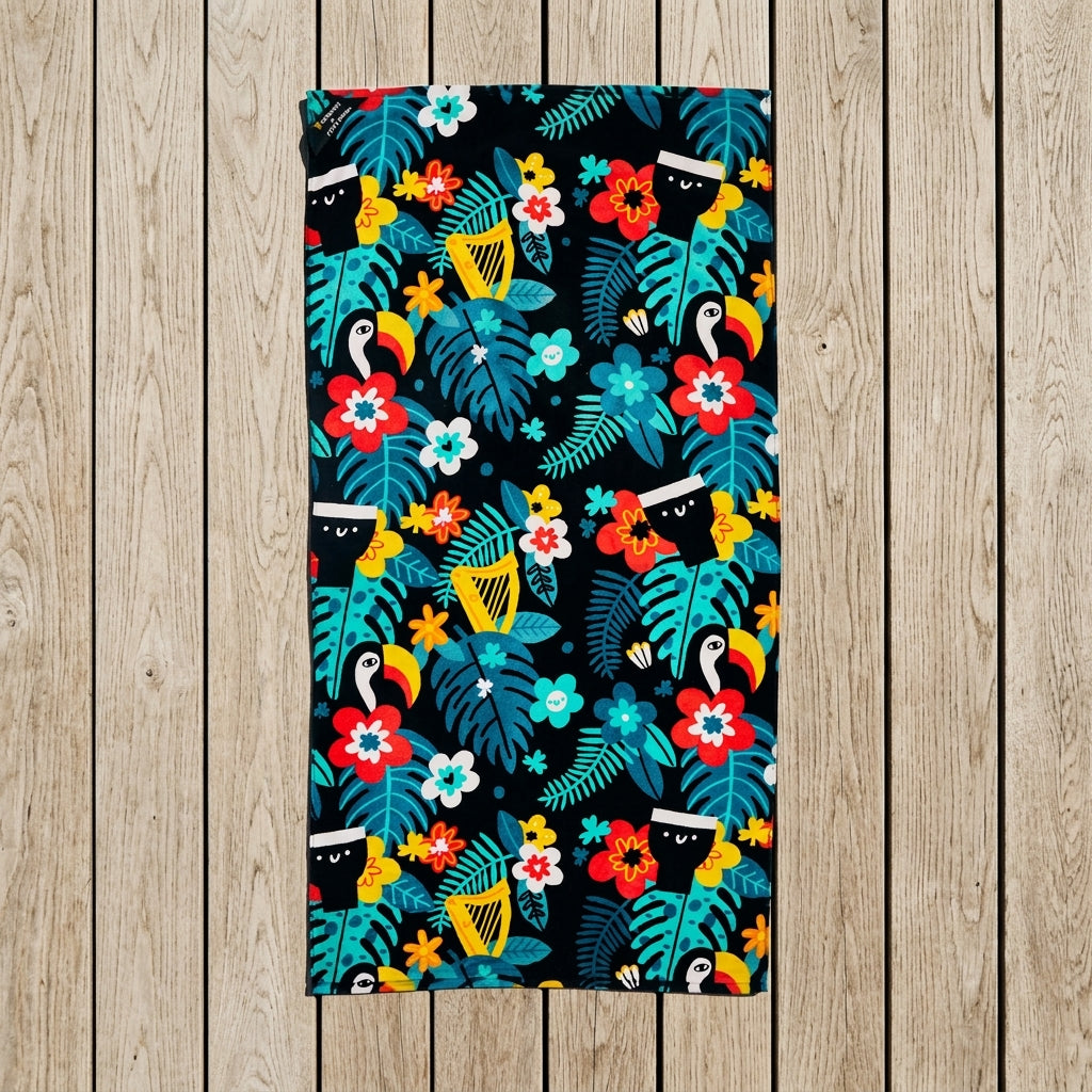 Guinness x Fatti Burke Hawaiian Beach Towel from Guinness Webstore US, featuring a colorful tropical pattern with toucans, leaves, and flowers, laid flat on a wooden floor.
