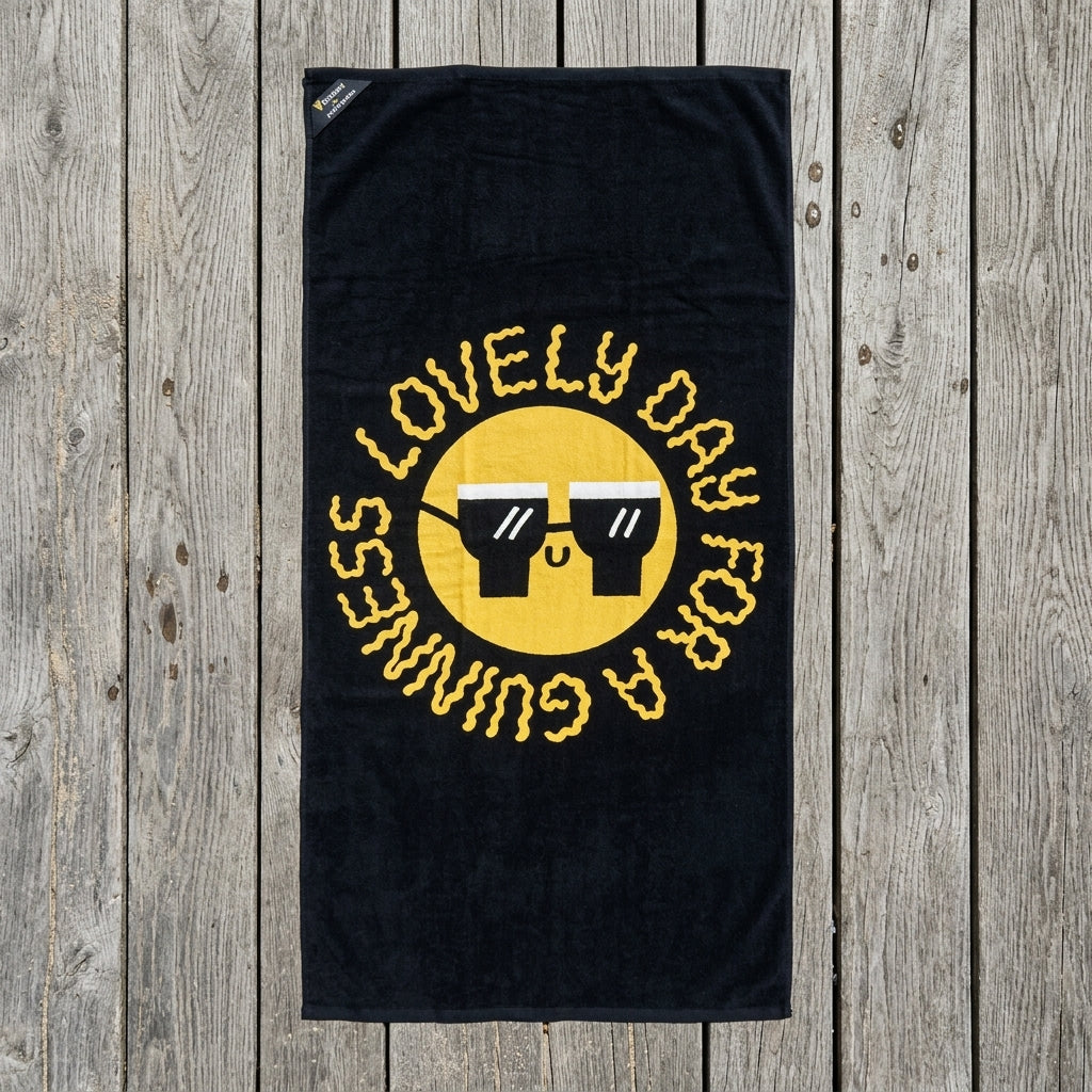 The Guinness x Fatti Burke Sun Beach Towel from Guinness Webstore US features a yellow smiley face with sunglasses and the words "LOVELY DAY FOR A GUINNESS" in a circular pattern on a black background, displayed on a wooden surface.