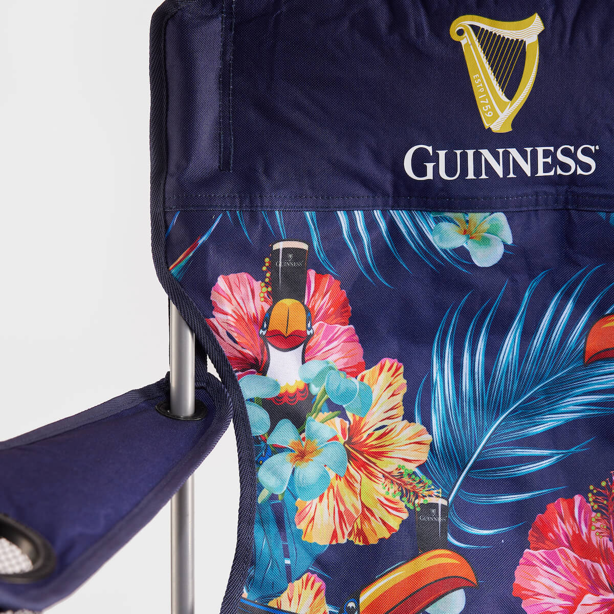 The Guinness Toucan Hawaiian Beach Chair by Guinness Webstore US is a blue, foldable beach essential featuring a vibrant tropical pattern of toucans, guitars, flowers, and the classic Guinness logo.