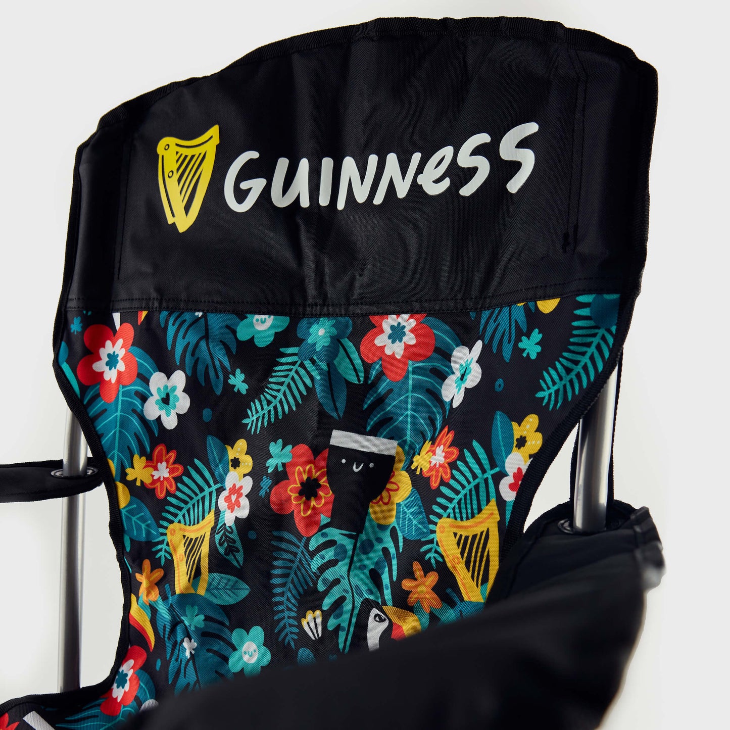 A close-up of the Guinness x Fatti Burke Hawaiian Camping Chair shows the Guinness logo on the black headrest and a vibrant floral backrest with toucan and pint glass illustrations, capturing the brand's iconic beach style.