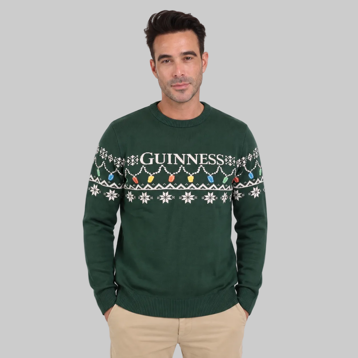 A man wearing the Guinness Holiday Pint Lights Sweater in green by Guinness Webstore US, which features “Guinness” text and a festive pattern, standing against a plain light background.