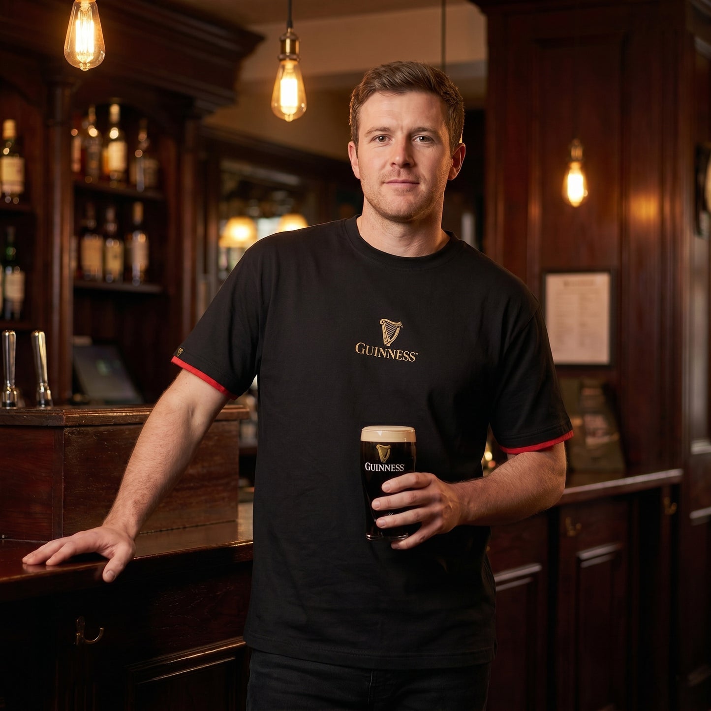 A man wearing a Guinness Irish Label Black Tee from Guinness Webstore US stands at a bar, holding a pint of Guinness. Warm lighting and bottles are visible in the background.