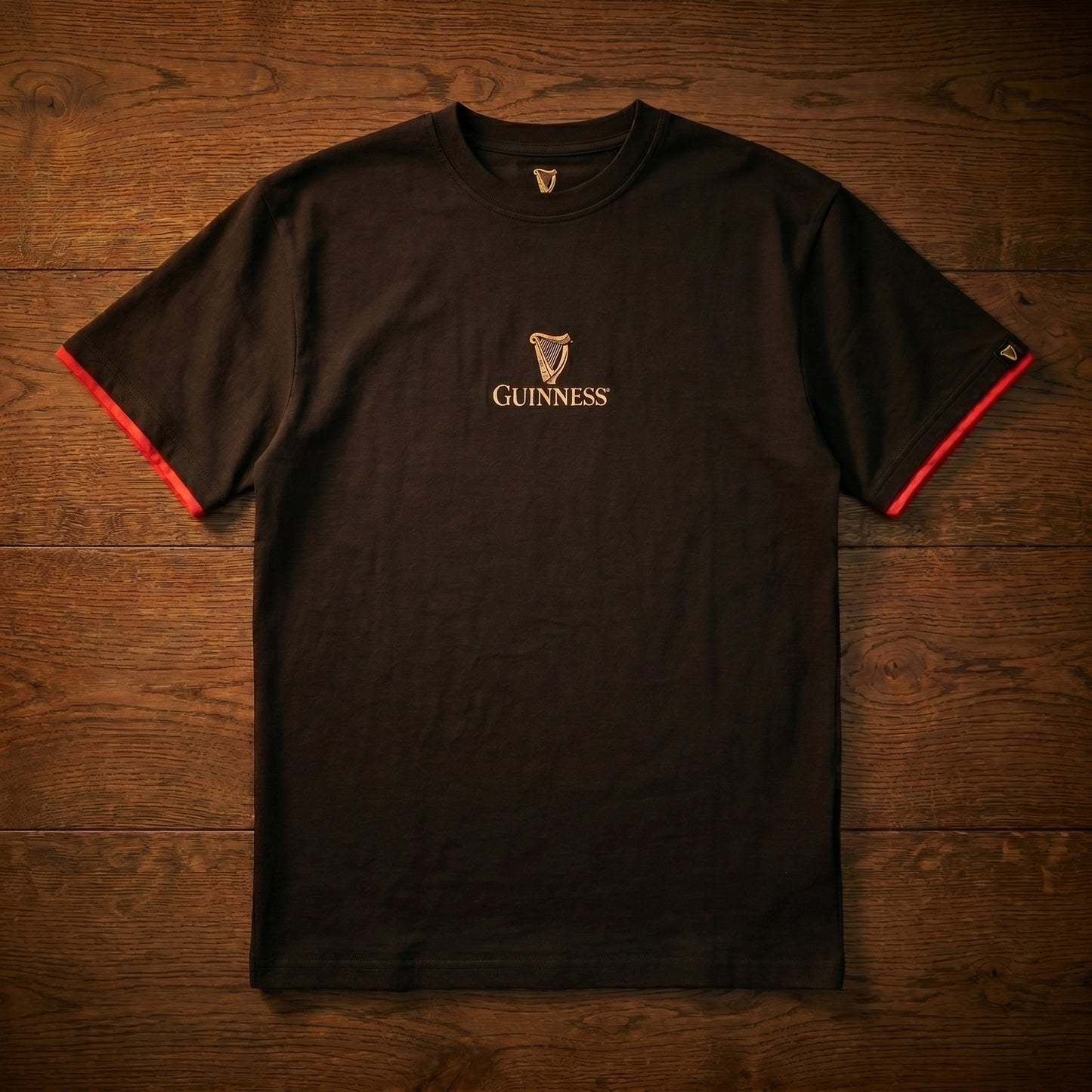 The Guinness Irish Label Black Tee by Guinness Webstore US features a small logo and harp on the chest, red-trimmed sleeves, and is displayed on a wooden surface—adding a stylish touch to this classic St. James's Gate Dublin shirt.