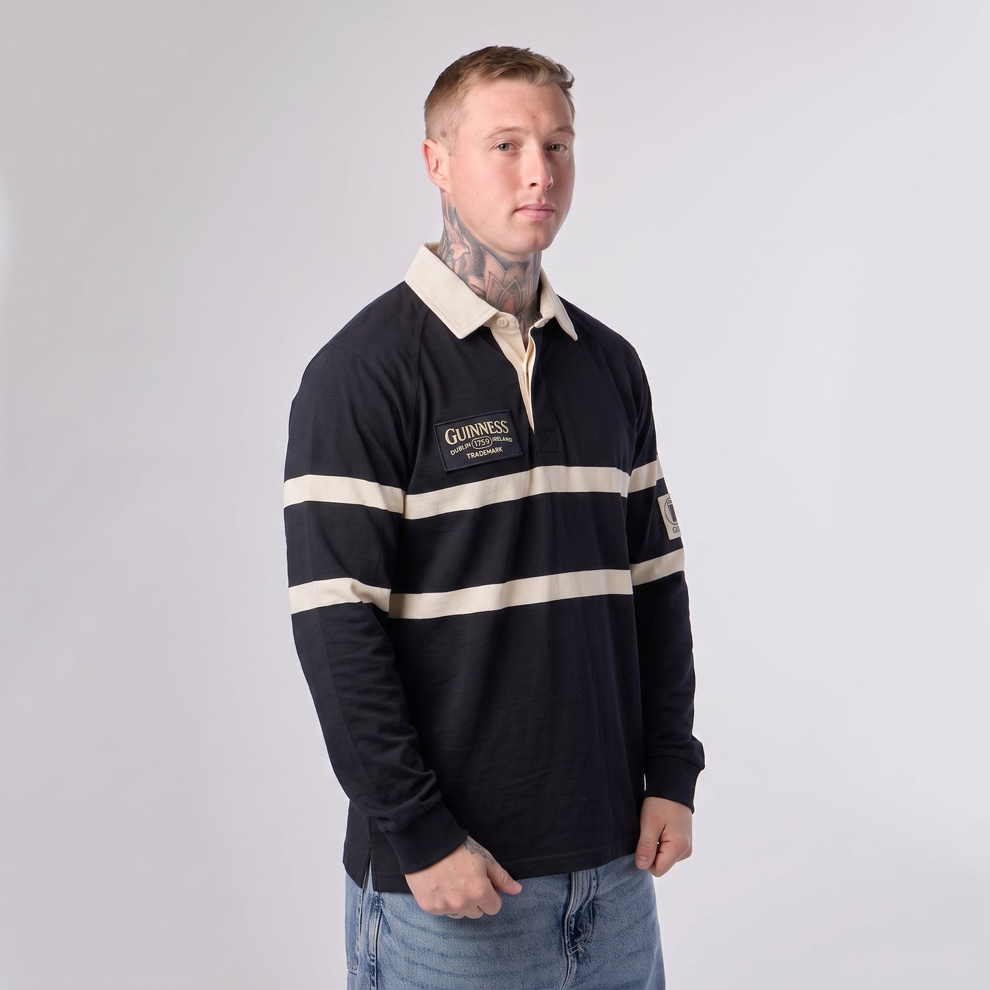 A man with tattoos stands against a plain light background wearing the Guinness Black Traditional Rugby Jersey by Guinness, crafted from 100% cotton in black and cream.