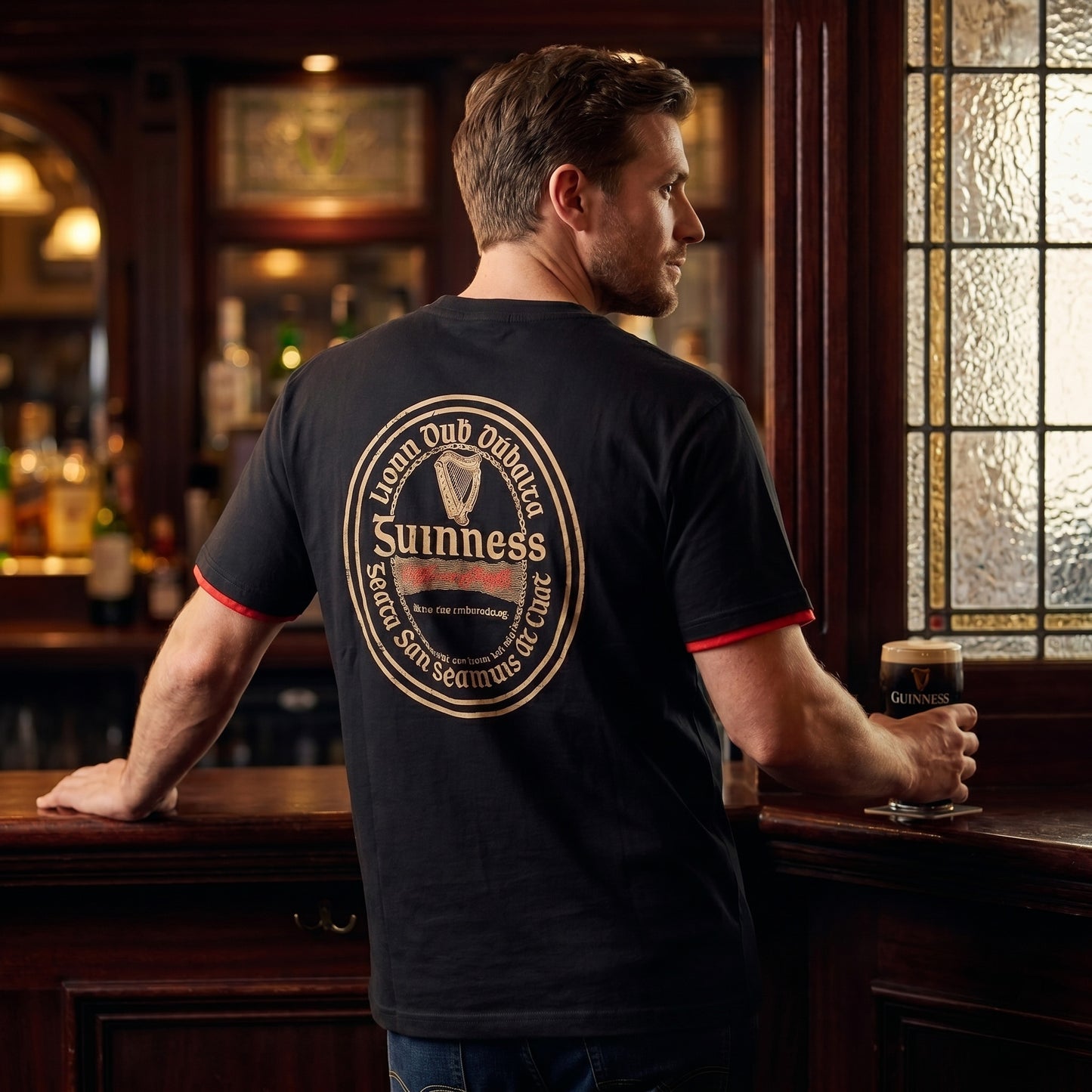 A man stands at a bar holding a pint of Guinness, wearing the Guinness Irish Label Black Tee from Guinness Webstore US, featuring a bold logo and Irish text on the back that celebrates St. James's Gate Dublin.