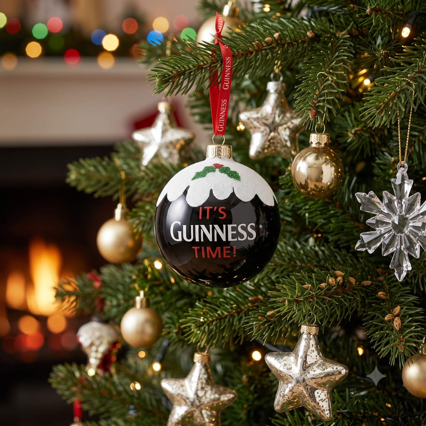 A Christmas tree adorned with gold and silver star ornaments, a clear snowflake, the Christmas Holiday Pudding Bauble by Guinness, and a round Guinness ornament displaying "It's Guinness Time!" stands near a fireplace with softly blurred holiday lights in the background.