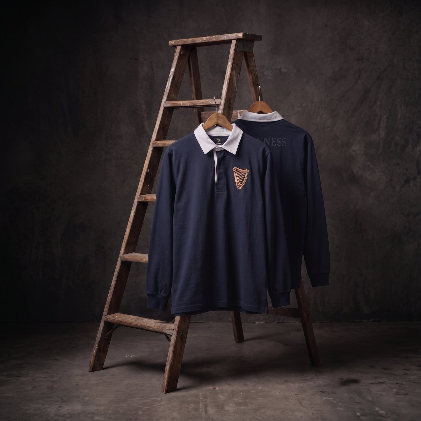 Two Guinness Navy Traditional Rugby Jerseys from the Guinness Webstore US, each featuring a classic white collar, are displayed on hangers in front of a wooden ladder and set against a dark, textured background.