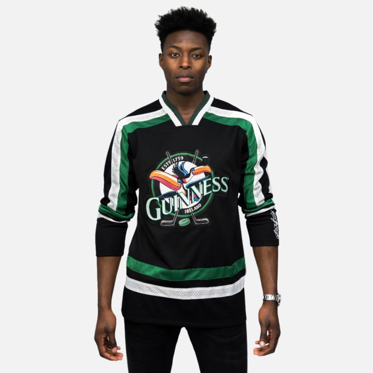 A man stands facing forward wearing the Guinness Toucan Hockey Jersey in black and green by Guinness, featuring white accents and a large Guinness logo with the iconic toucan on the front.
