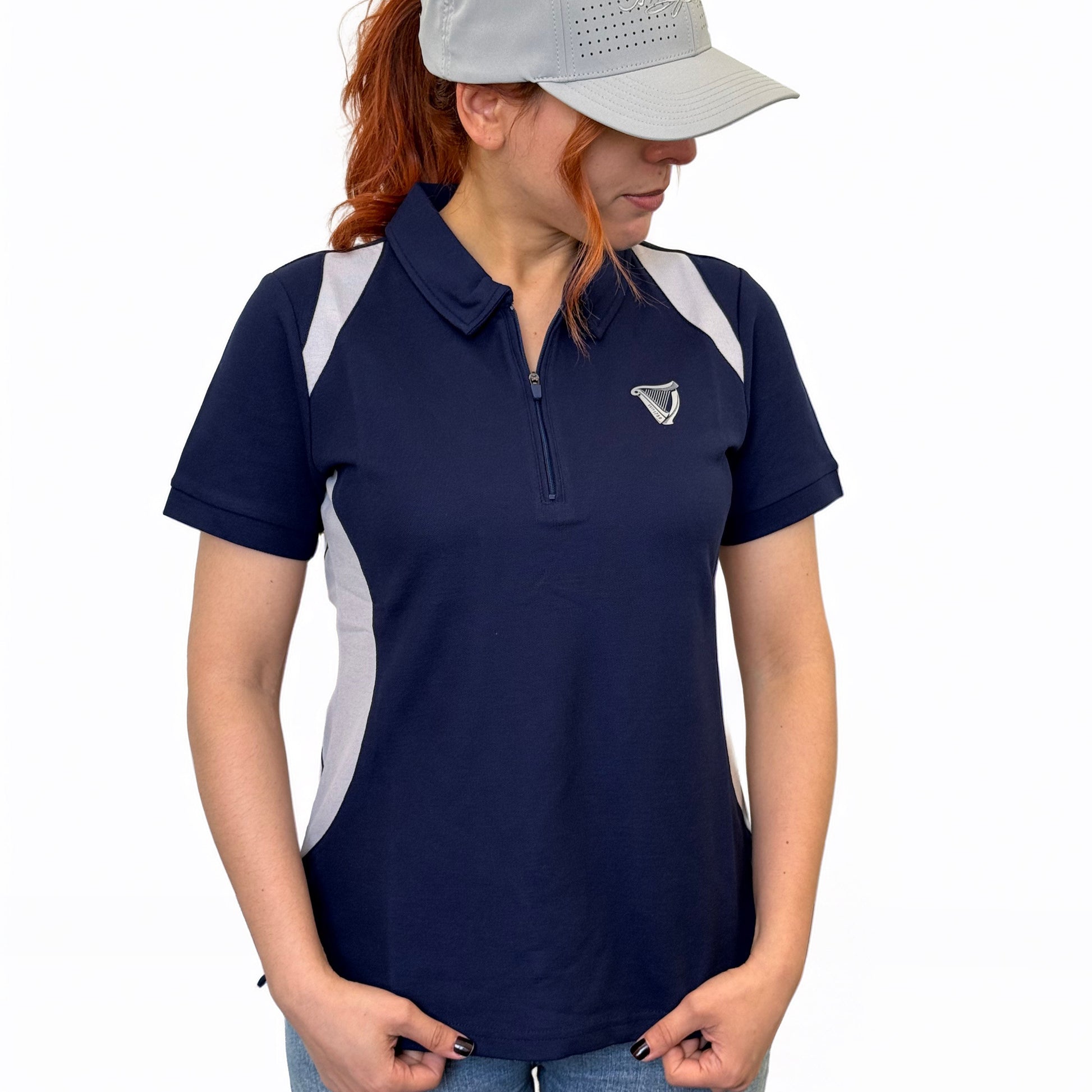A woman in the Guinness Webstore US Ladies Performance Polo - Blue stands with hands in pockets and looks down, pairing it with a light gray baseball cap.