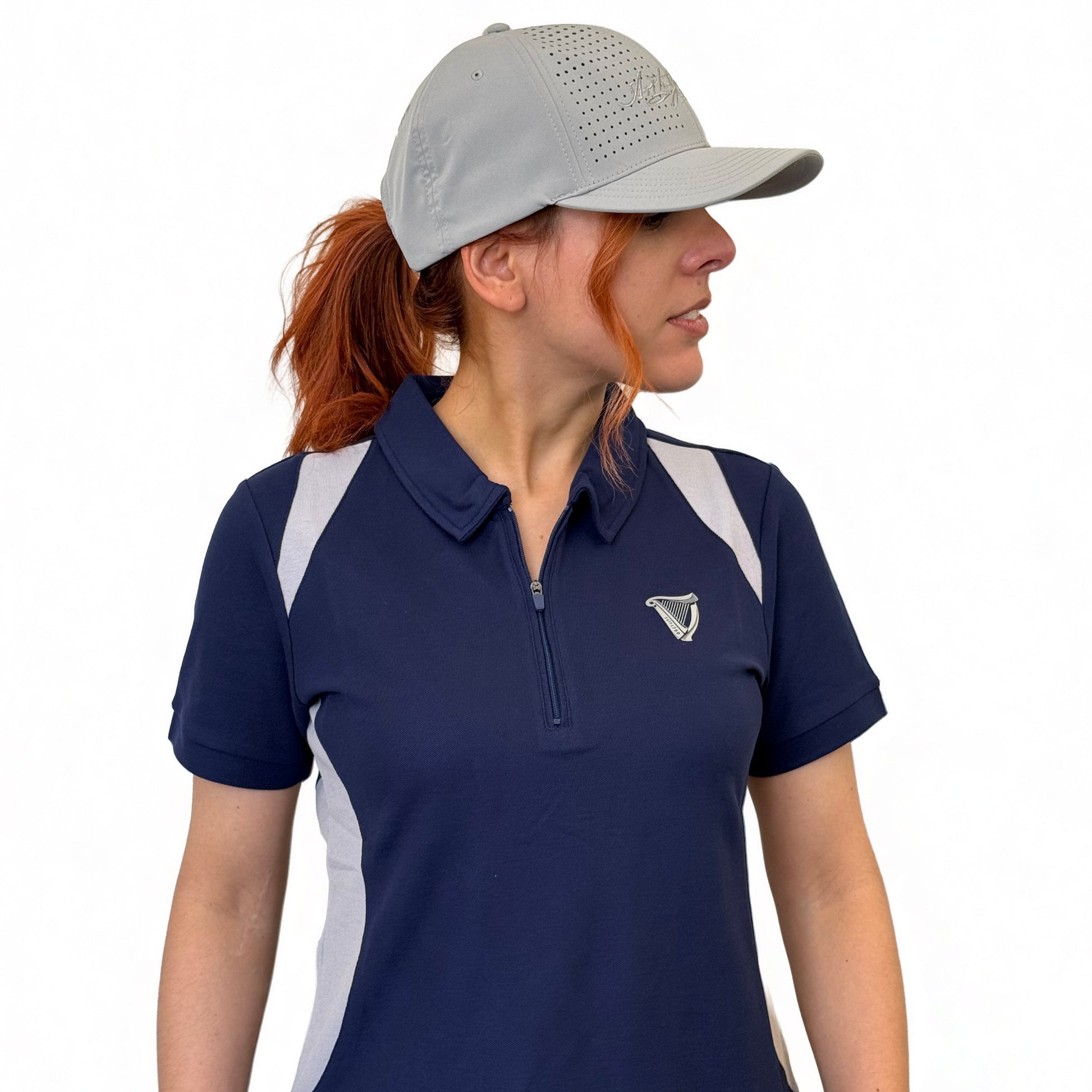A woman in a gray cap, blue Guinness Webstore US Ladies Performance Polo, and blue jeans stands against a white background, looking to her left.