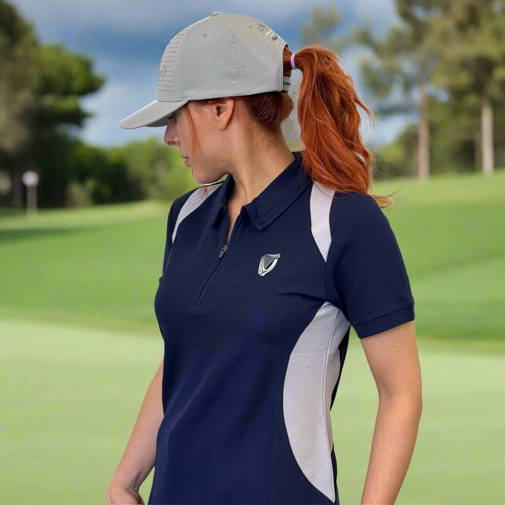 A woman wearing a gray cap, light blue jeans, and the Guinness Webstore US Ladies Performance Polo - Blue with a small logo stands against a white background, looking to her left.